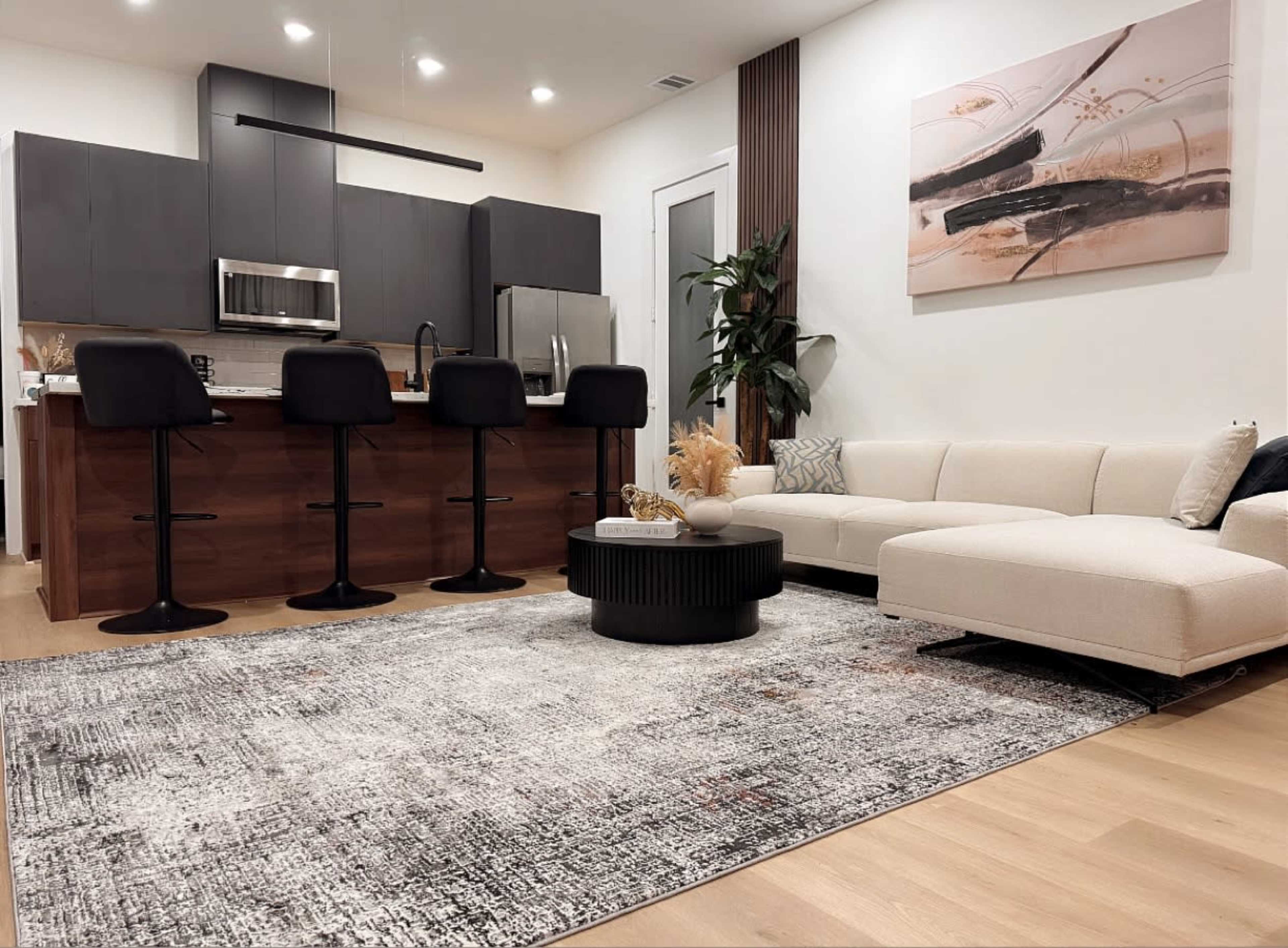 The image shows a modern living space featuring a sleek kitchen with bar stools, a sectional sofa, and a decorative rug.