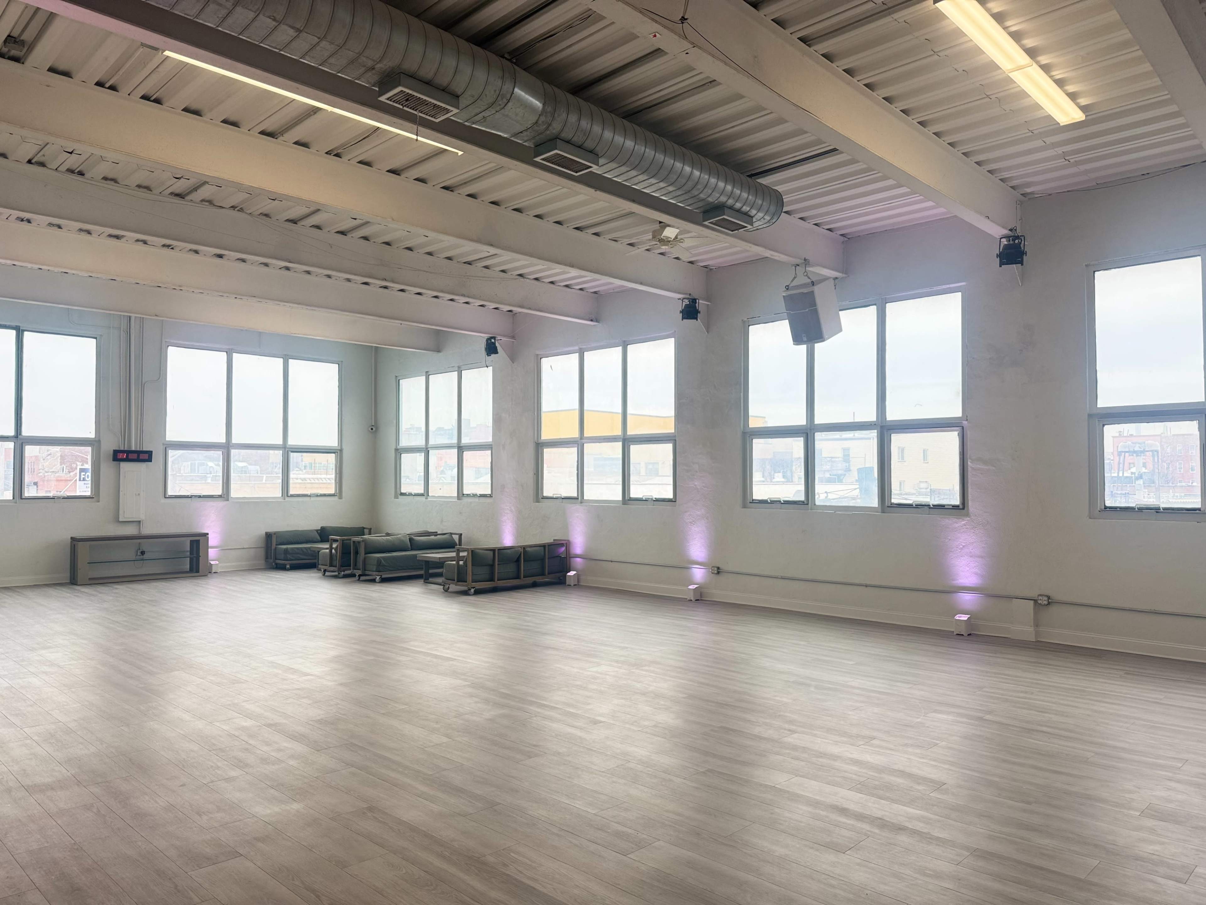 The image shows a spacious interior room with large windows, minimal furniture, and purple lighting along the walls.