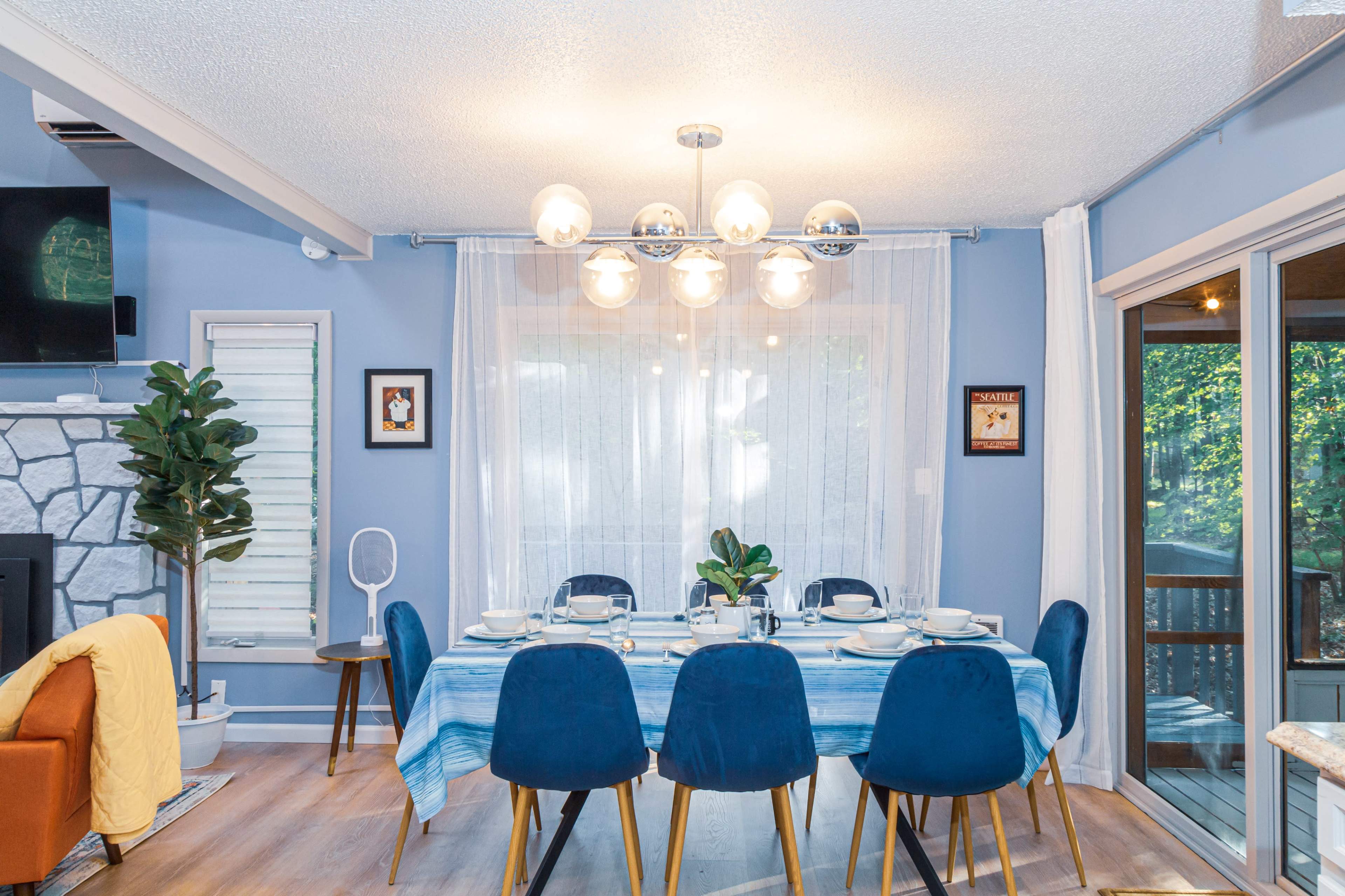 A dining area features a table set for six, with blue chairs, a decorative light fixture, and large windows offering a view of the outdoors.