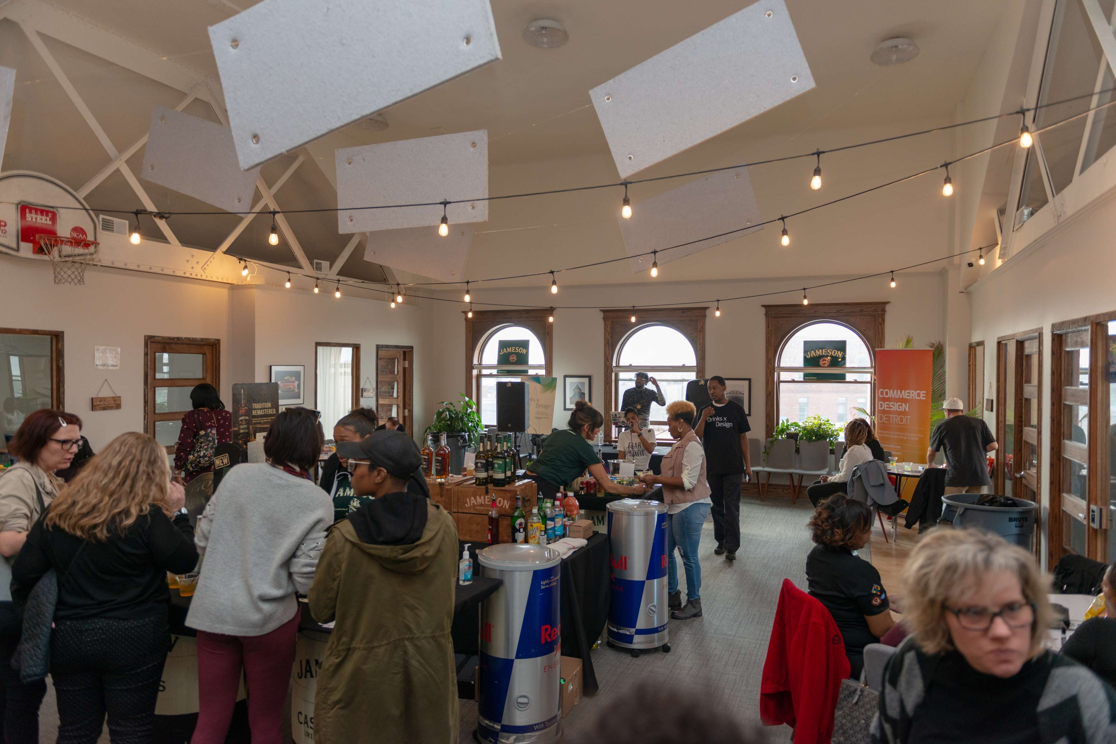 A busy event space with people mingling around several beverage stations and tables, under warm string lights and a basketball hoop on the wall.