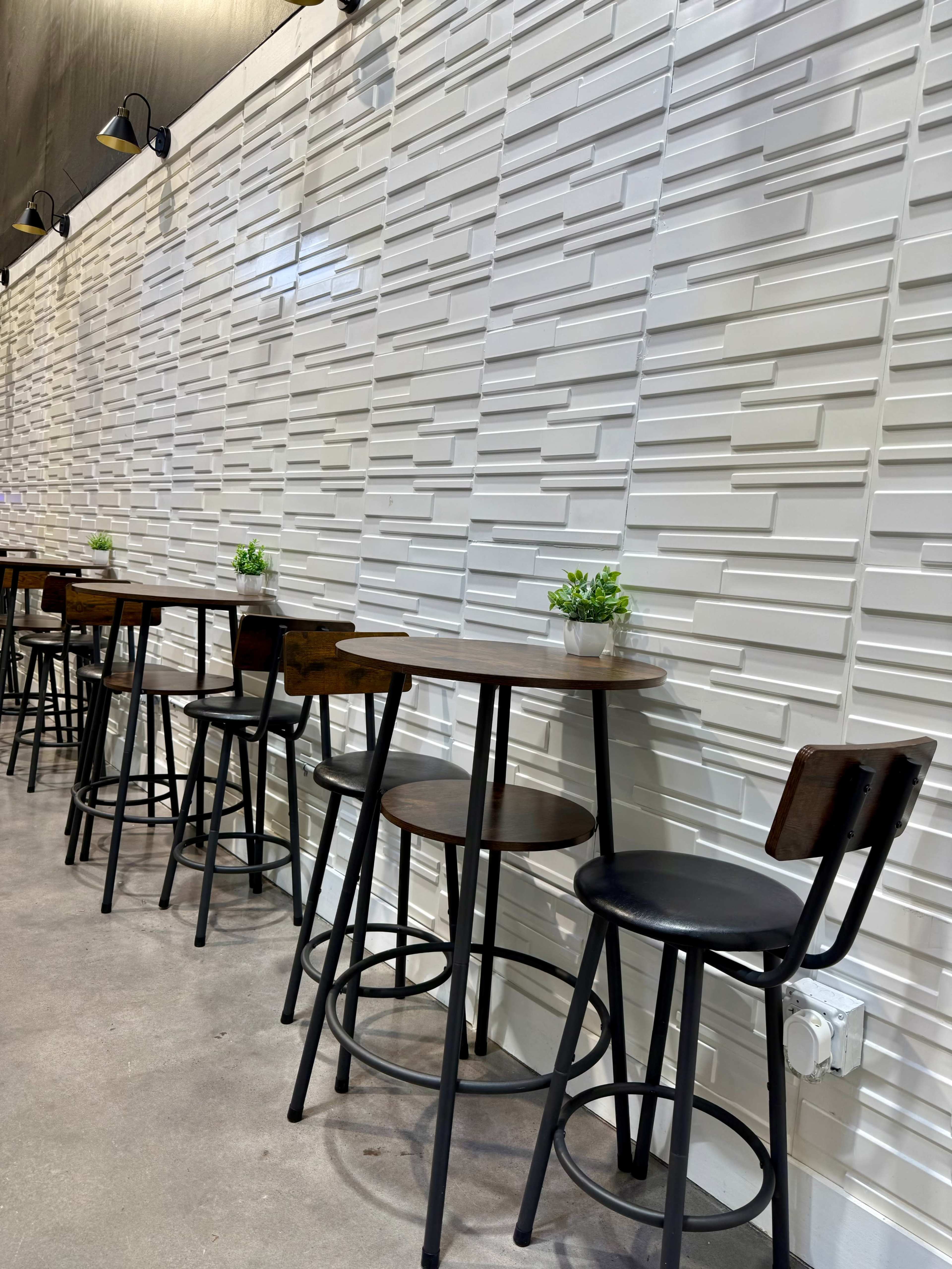 The image shows a series of tall tables and chairs arranged against a textured white wall in a modern café setting.