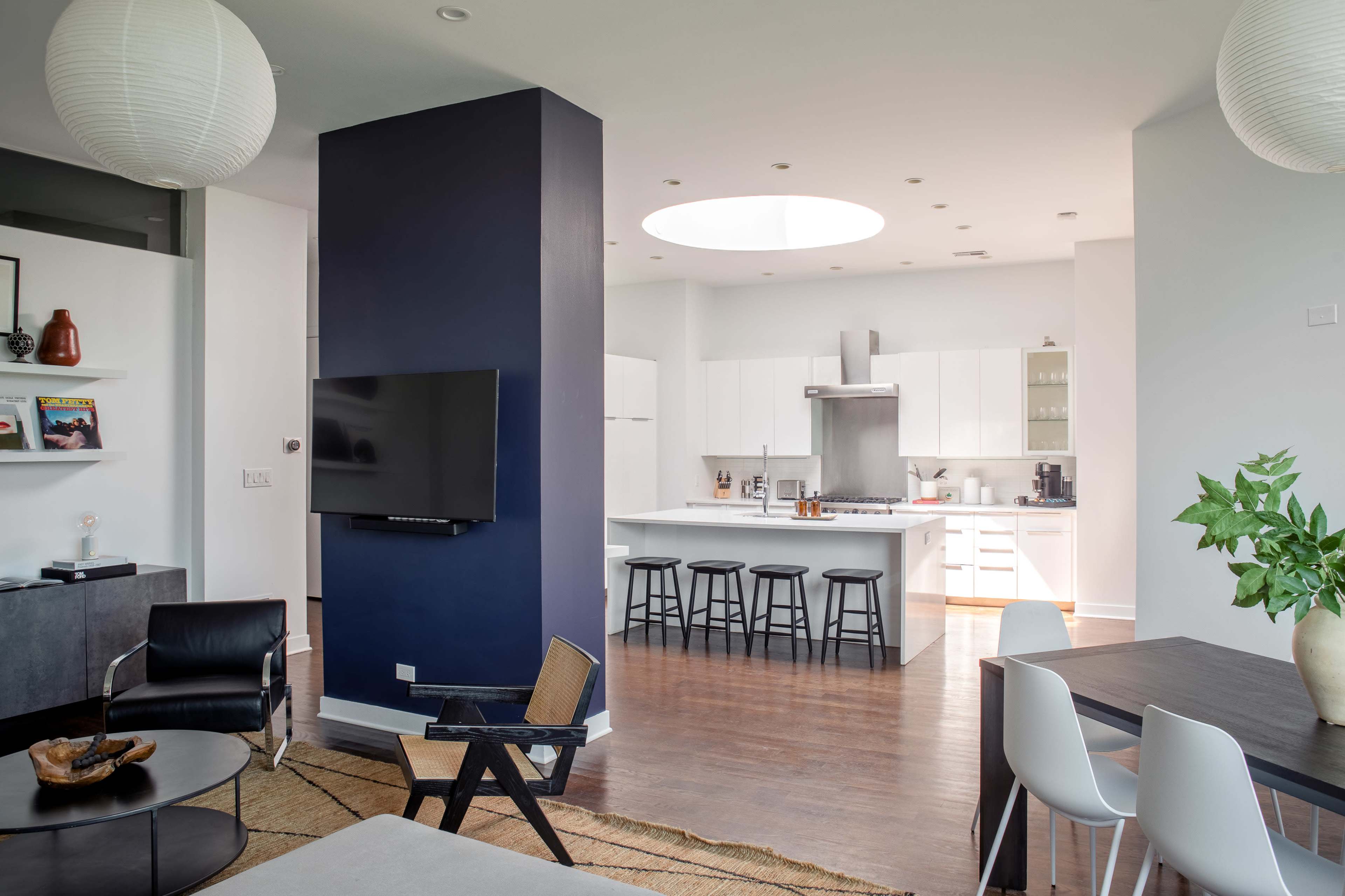 A modern, open-plan living space featuring a kitchen with white cabinets, a dining area with a wooden table, and a living area with a black chair and coffee table.
