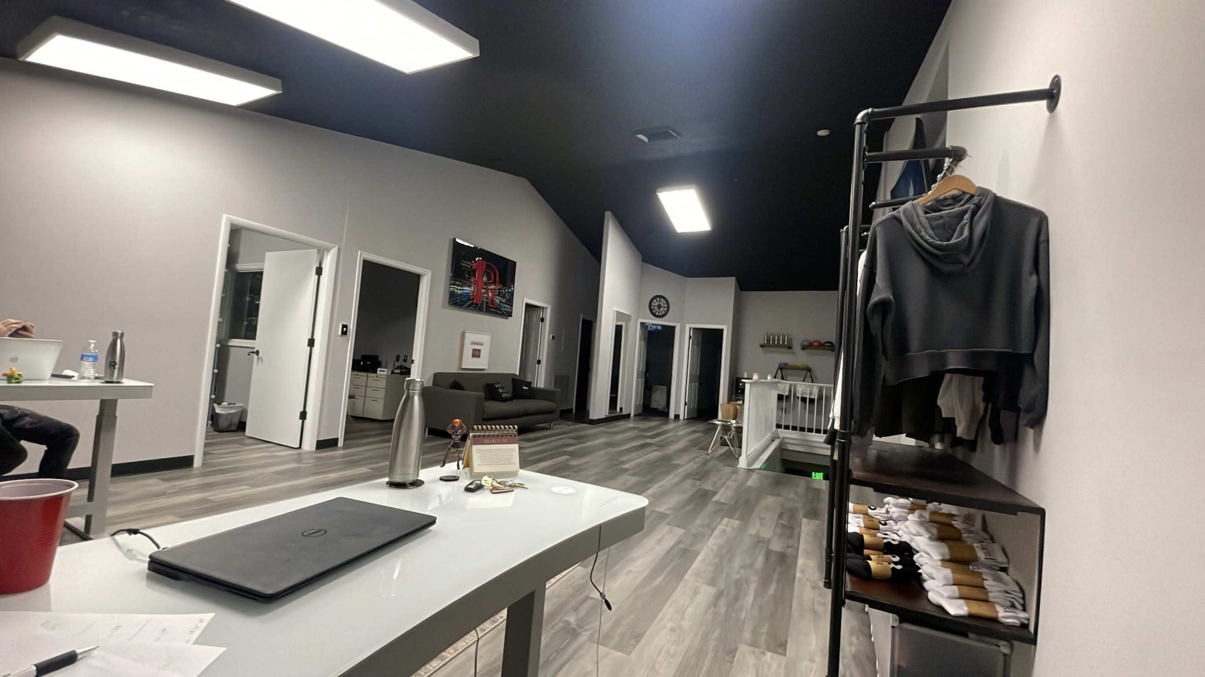 A modern interior of a workspace or studio with gray walls, wooden flooring, and several distinct areas including a lounge with a sofa and a clothing rack.