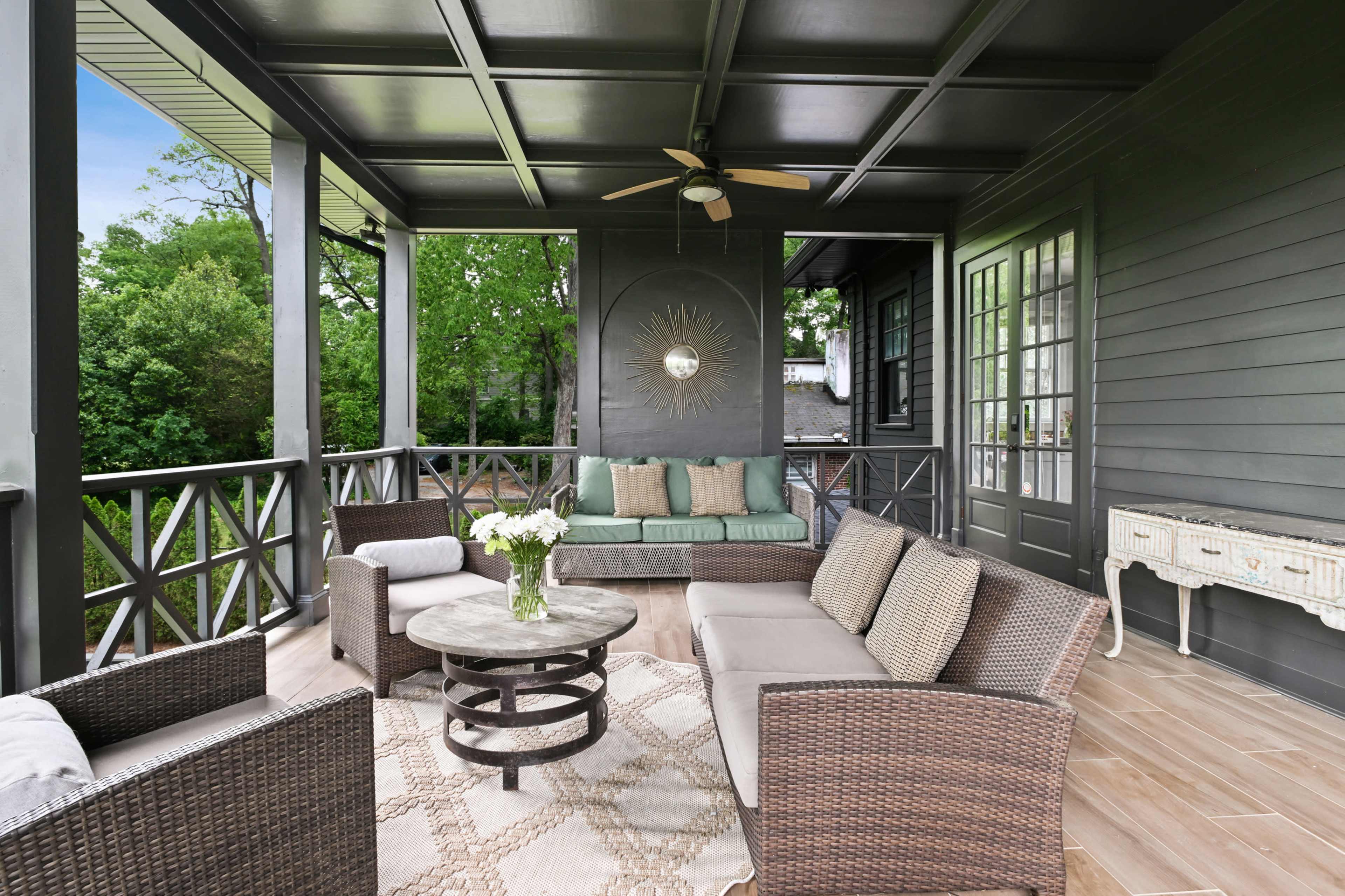 A modern porch features a round table with a floral arrangement, surrounded by wicker seating and a wall-mounted sunburst mirror.