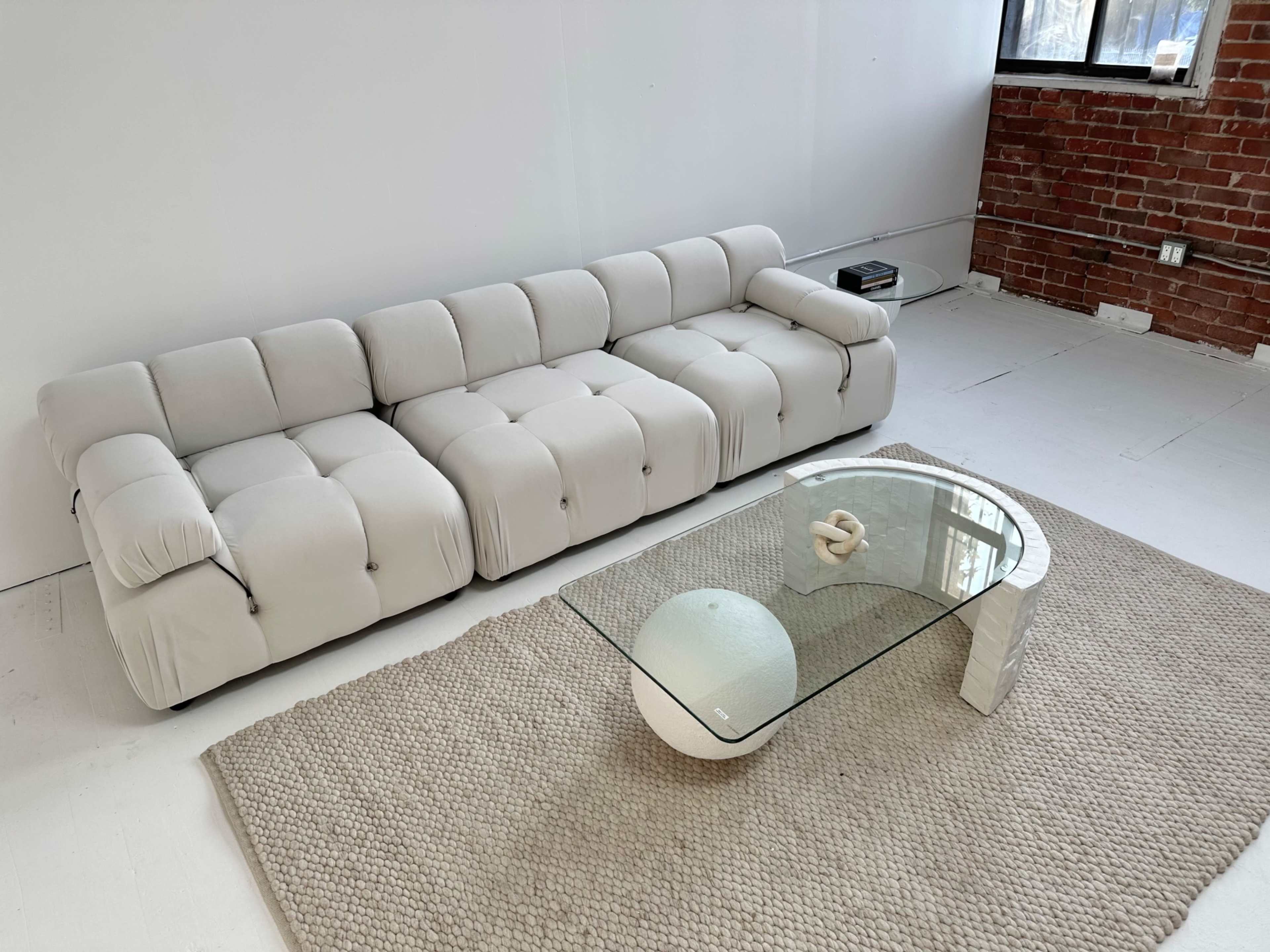 A modern white sofa with tufted upholstery, accompanied by a glass coffee table and a round decorative object, set on a textured rug in a room with exposed brick walls.