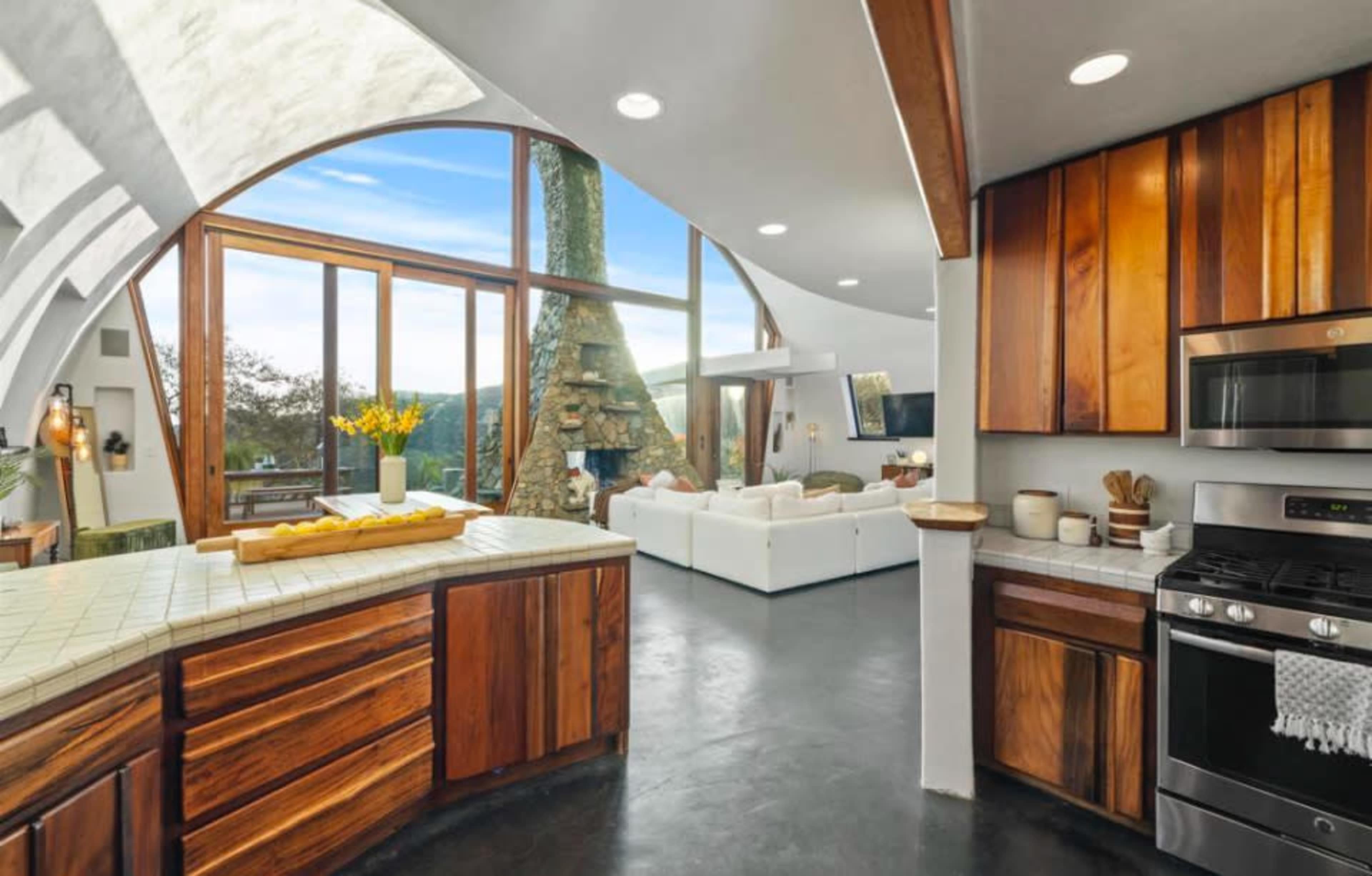 The image shows a modern kitchen featuring wooden cabinetry, a gas stove, and an open layout that leads into a living area with large windows and a view of the outdoors.