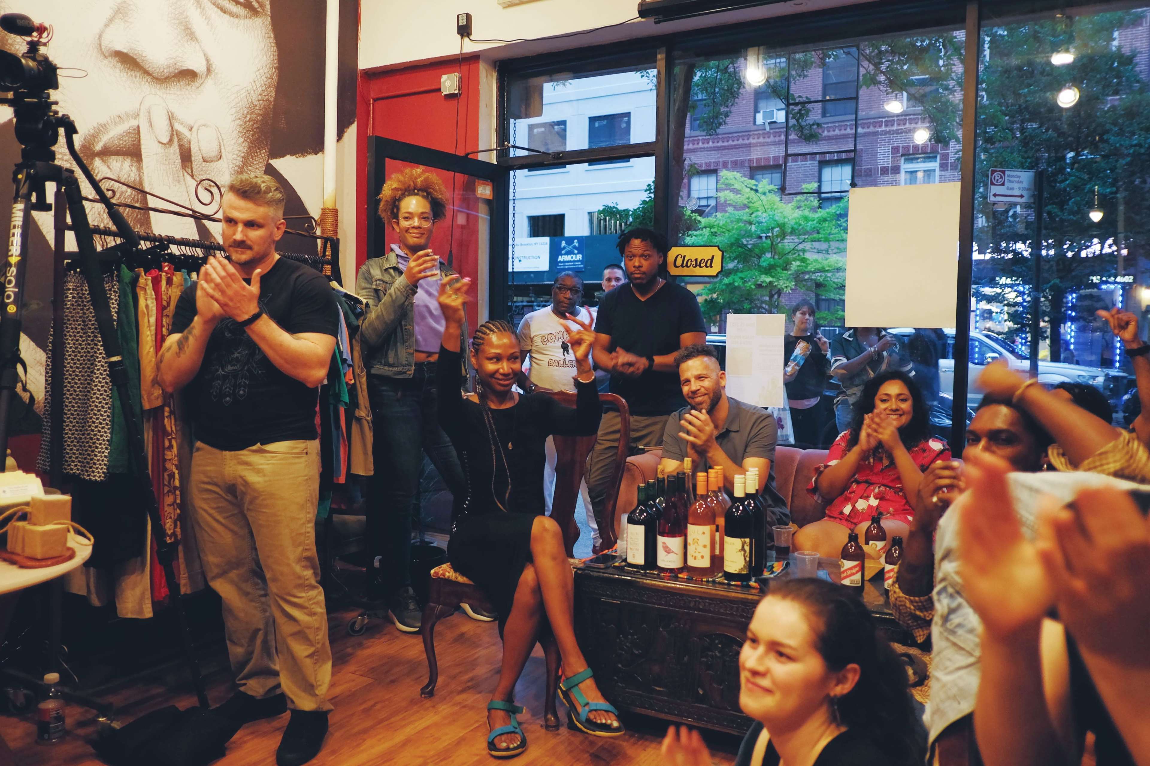 A group of people is gathered inside a store, clapping and sitting around a table with bottles of wine, while others stand nearby in front of a large window.