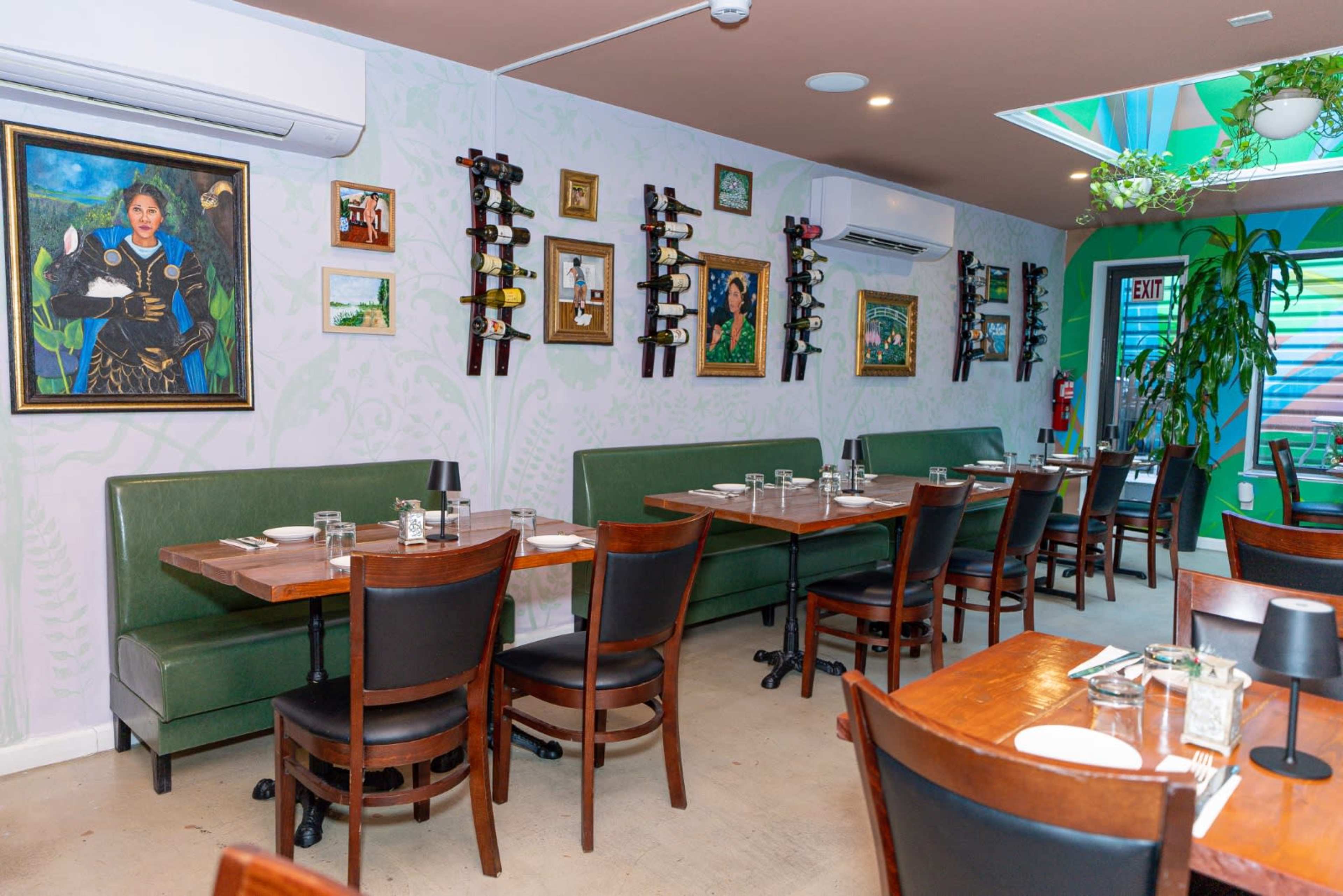 The image shows an indoor restaurant setup with green booths and wooden tables arranged neatly, adorned with framed artworks and wine racks on the walls.