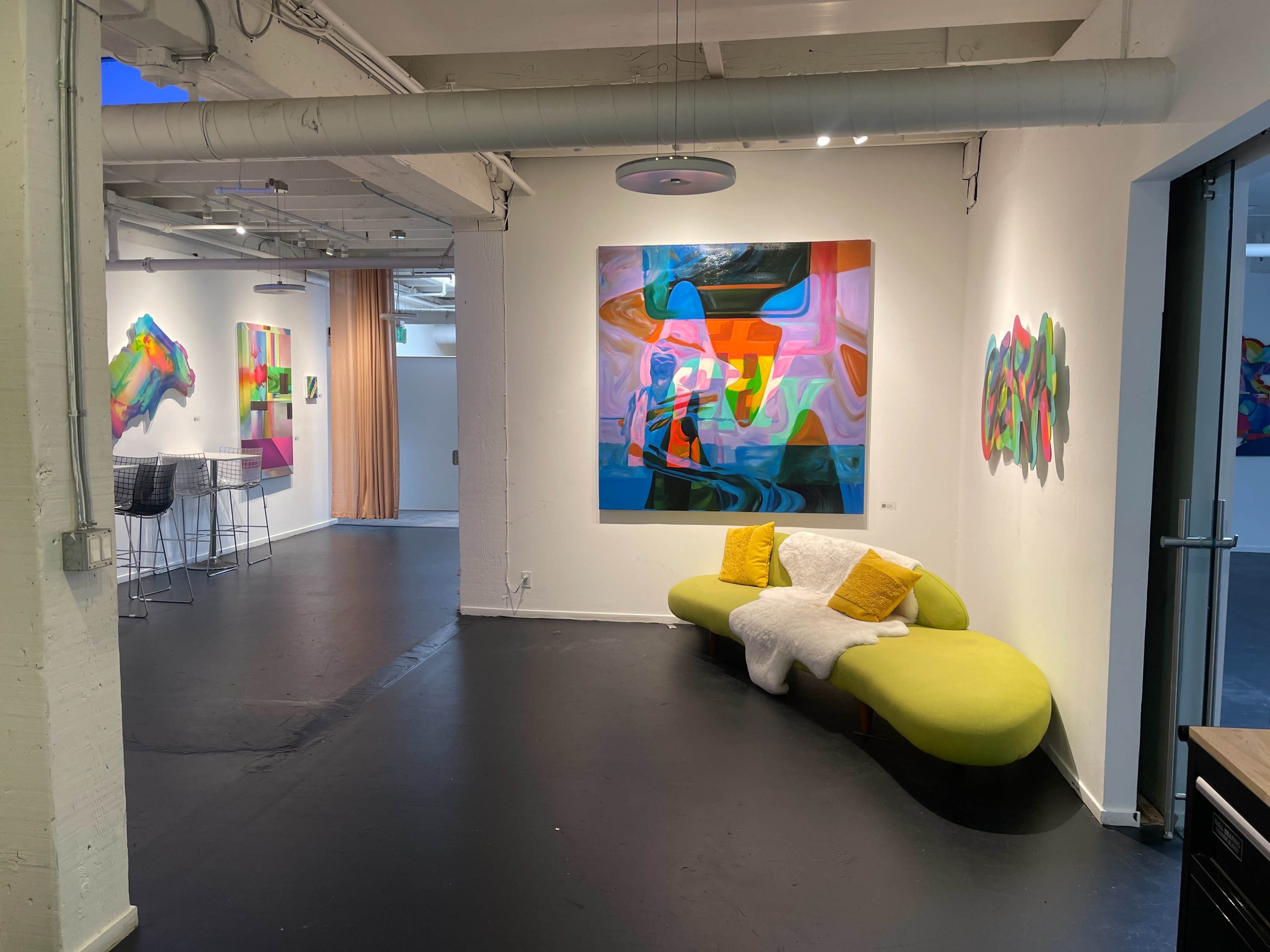 The scene features an art gallery with colorful abstract paintings on the walls, a green curved sofa with decorative cushions, and a seating area in the background.