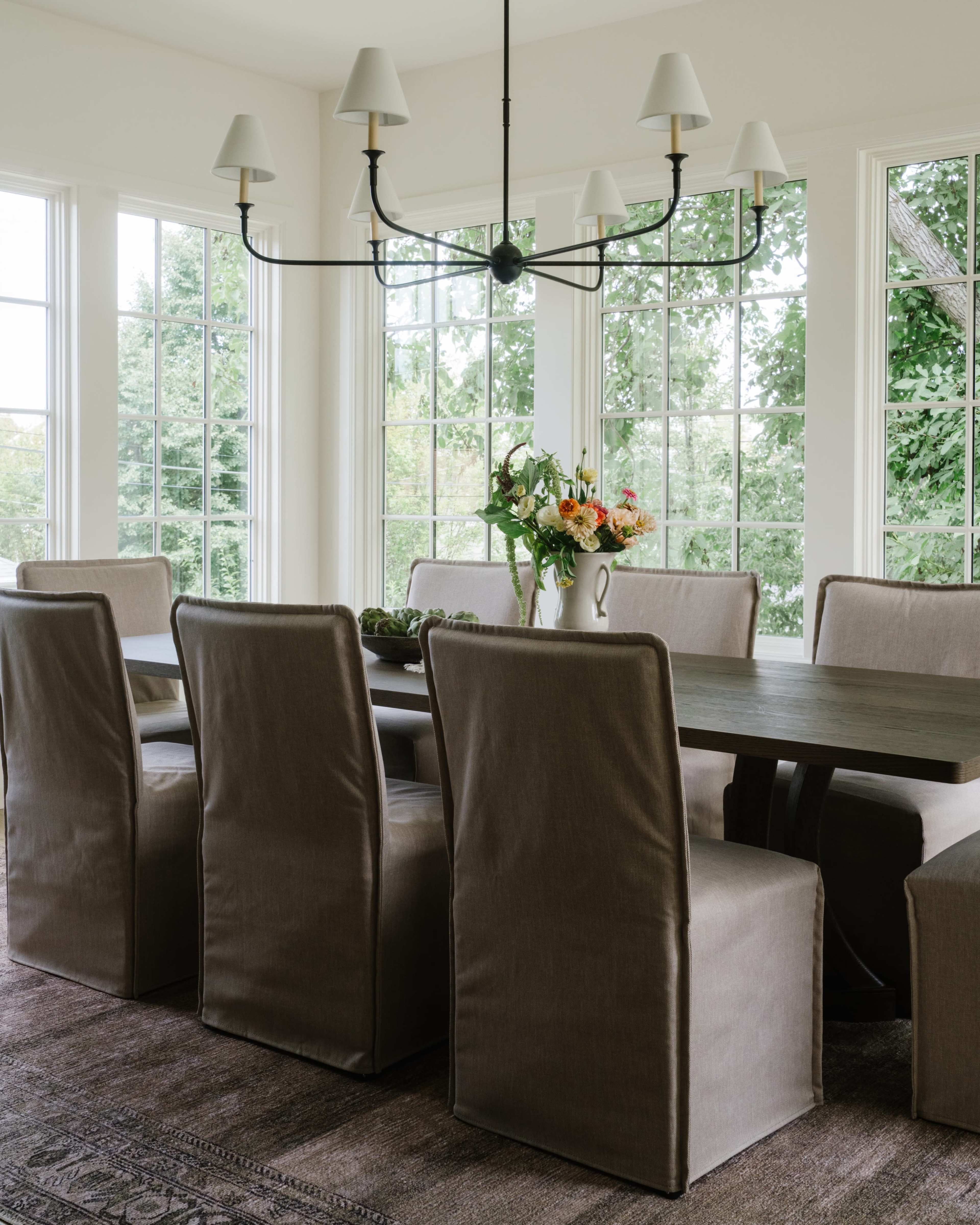 A dining room features a long wooden table surrounded by upholstered chairs, with a chandelier overhead and large windows allowing natural light to fill the space.