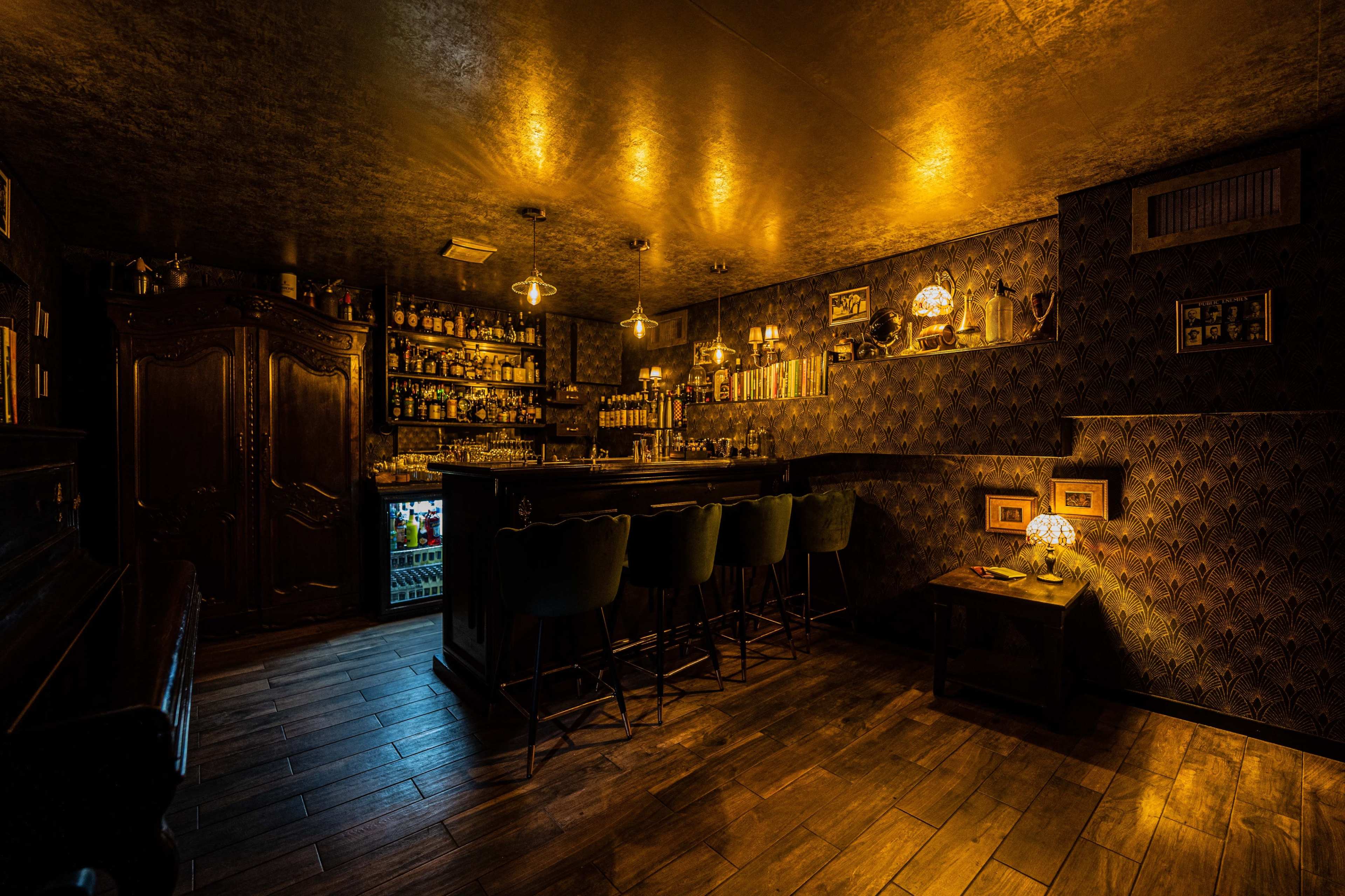 A dimly lit bar interior featuring a dark wooden counter, high-backed stools, and shelves filled with various bottles against a textured wall.