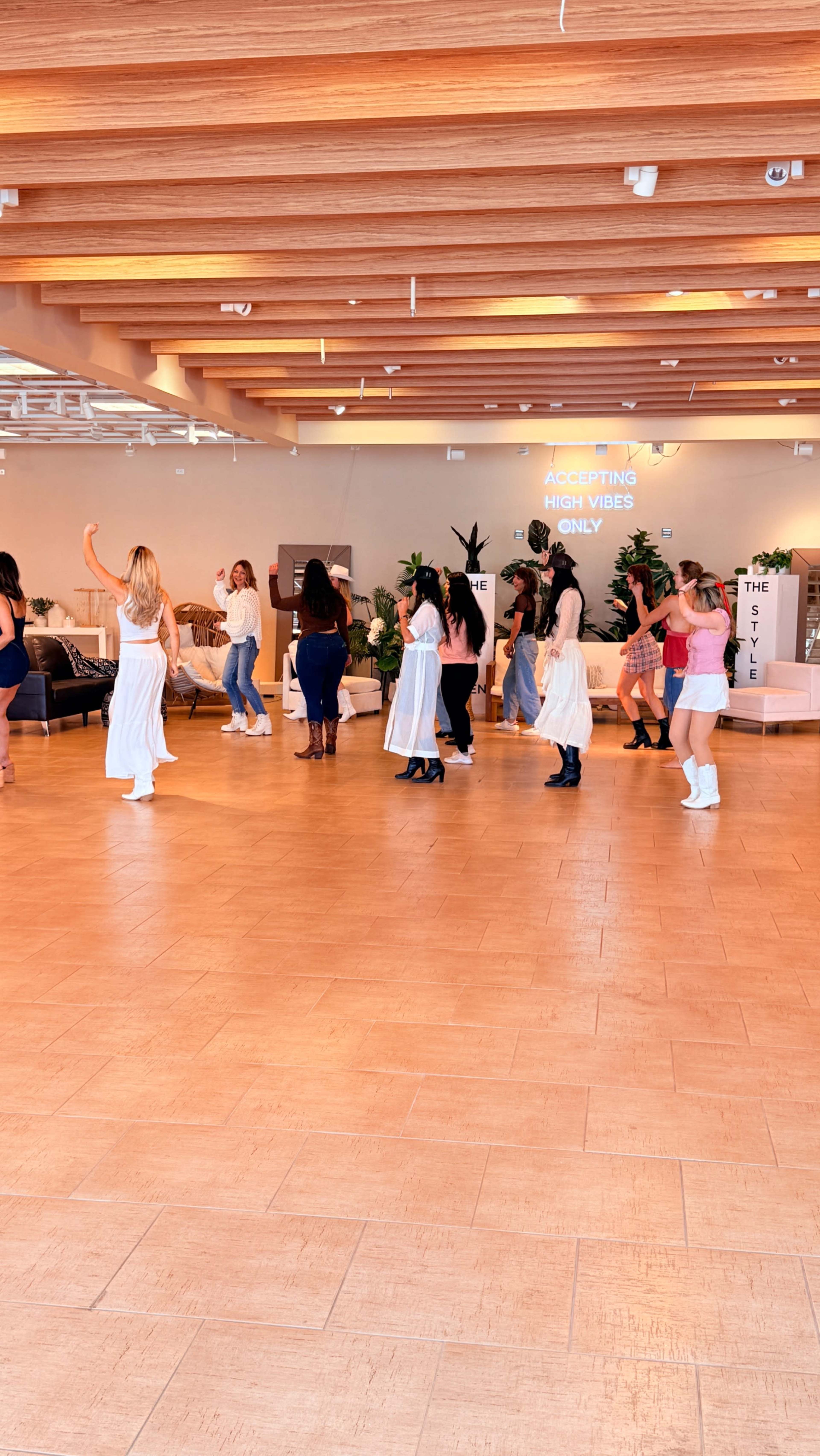 A group of women dressed in various outfits are dancing in a spacious room with wood paneling and plants, while a sign on the wall reads "ACCEPTING HIGH VIBES ONLY."