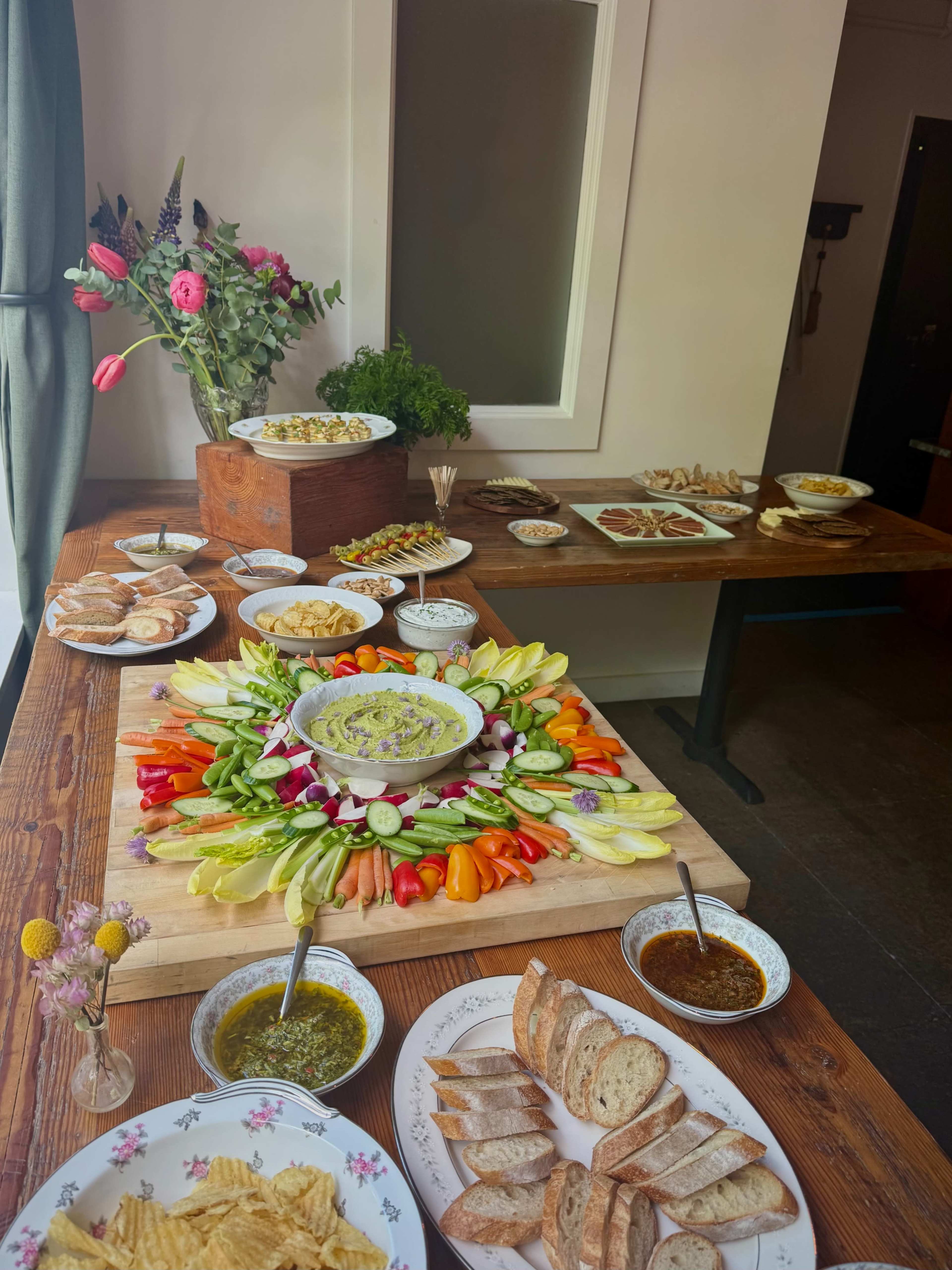 A large wooden table is adorned with an array of colorful vegetables arranged around a bowl of dip, alongside various plates of snacks, spreads, and bread.