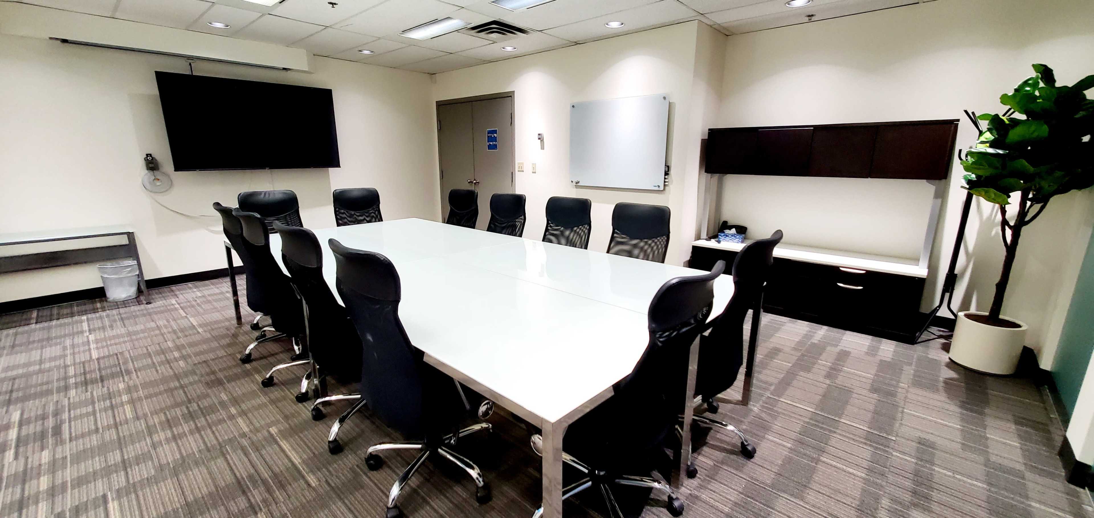 A modern conference room featuring a long white table surrounded by black ergonomic chairs, with a whiteboard and two screens mounted on opposite walls.