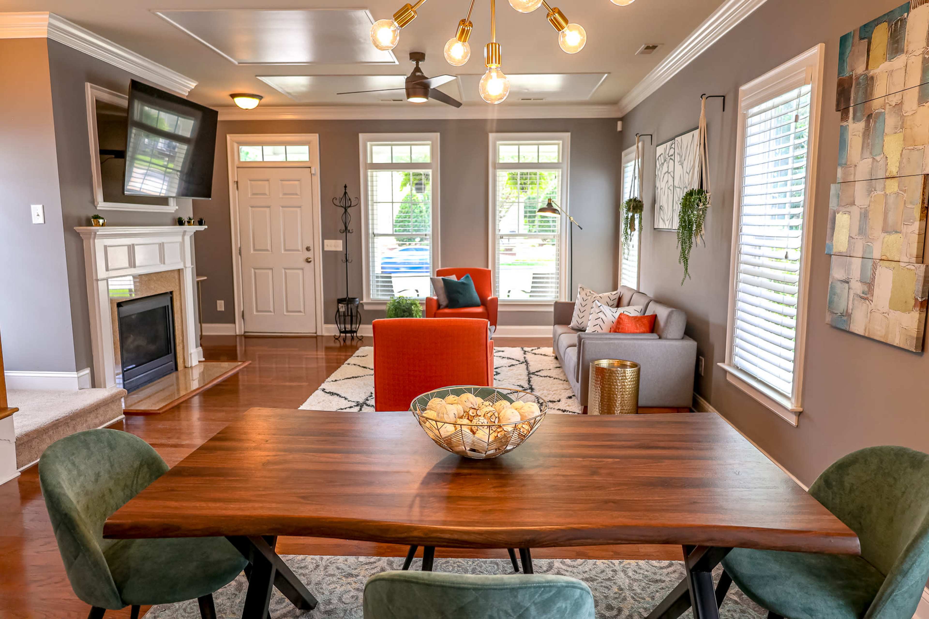 A modern living room features a large dining table, a gray sofa, an orange accent chair, a fireplace, and large windows allowing natural light.