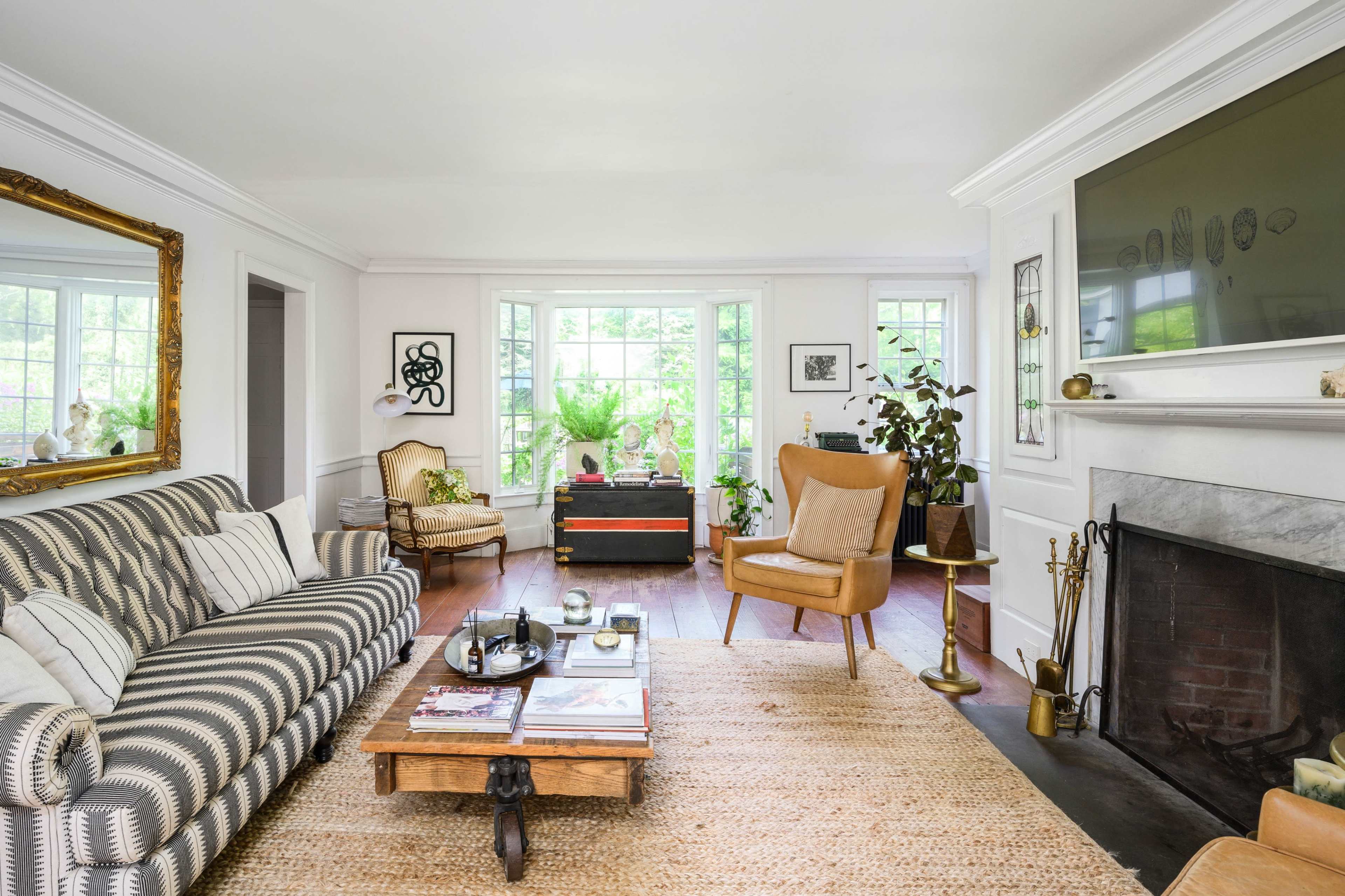 The living room features a striped sofa, a wooden coffee table, and a fireplace, with large windows allowing natural light and indoor plants enhancing the decor.