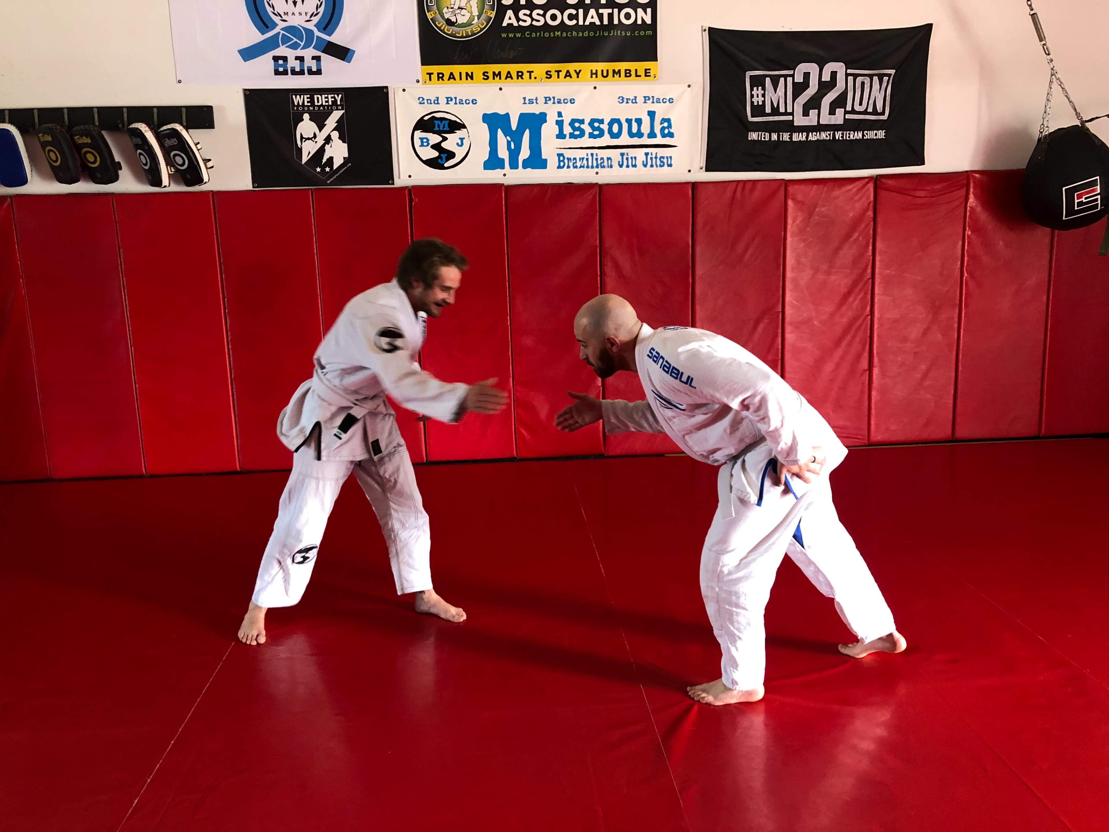 Two individuals in Brazilian Jiu-Jitsu gi uniforms are shaking hands on a red mat inside a training facility.