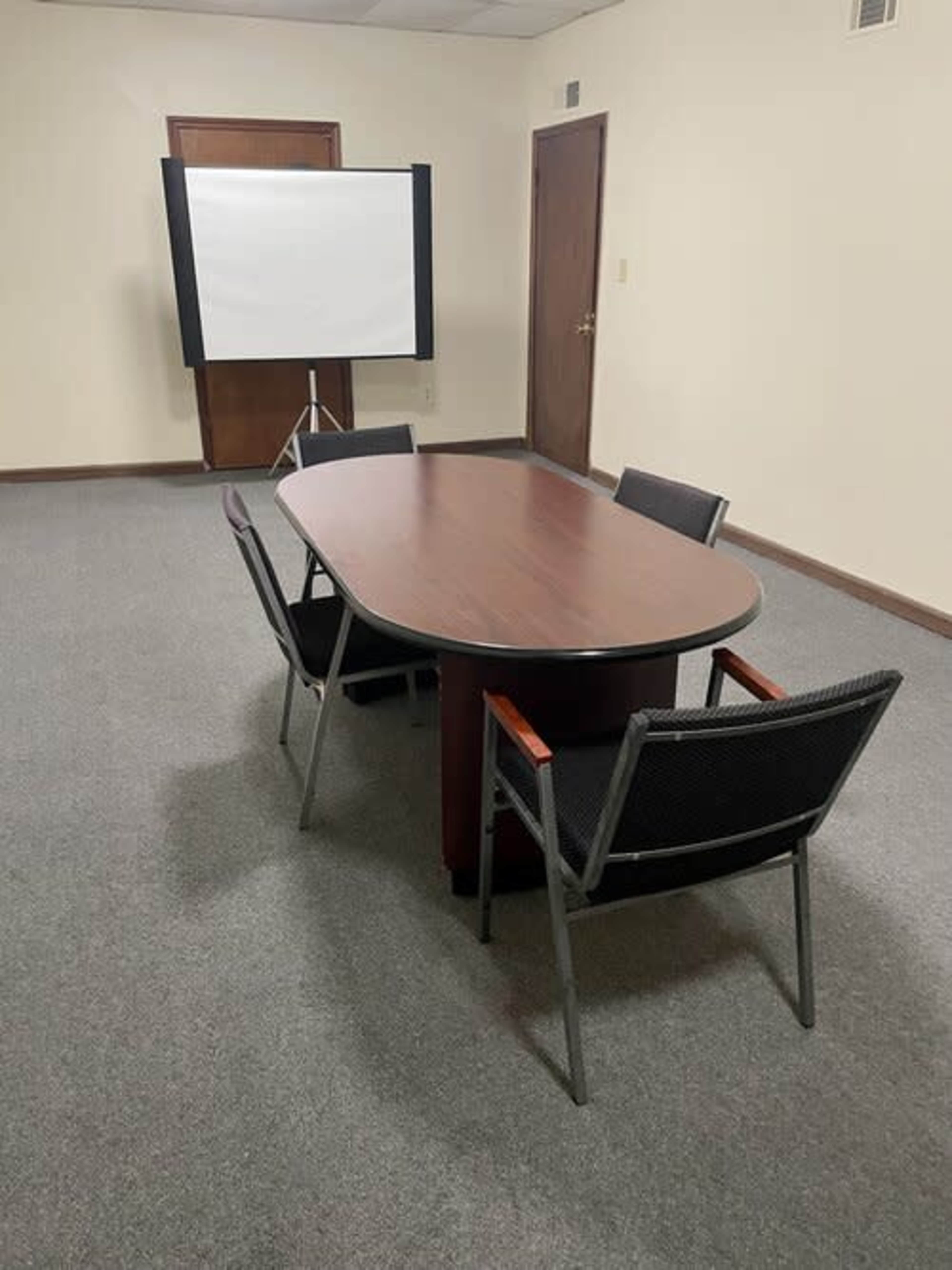 A rectangular table with four chairs is positioned in a sparsely furnished meeting room with a projection screen at one end.