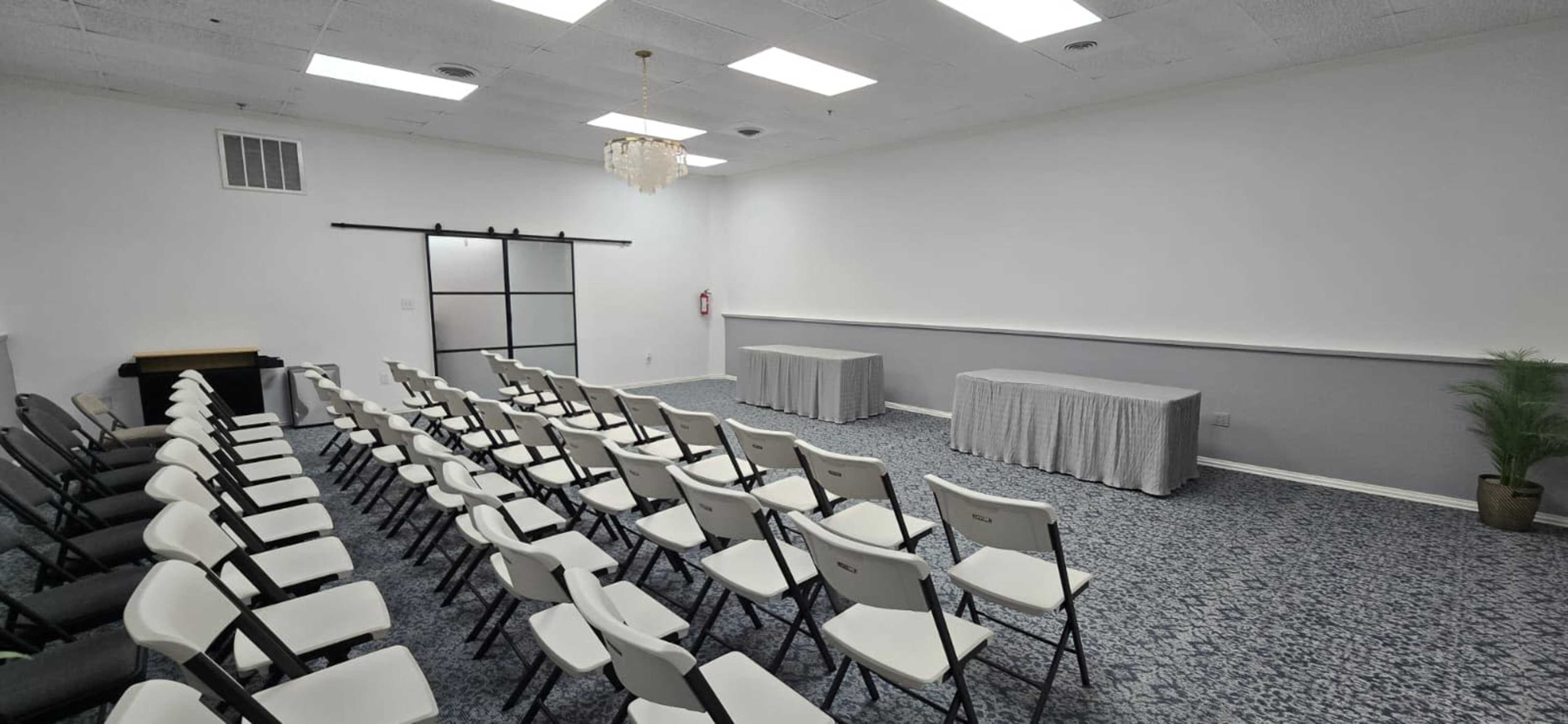 The image shows a conference room with several rows of white folding chairs facing two tables covered with gray tablecloths, all set against a plain white wall.