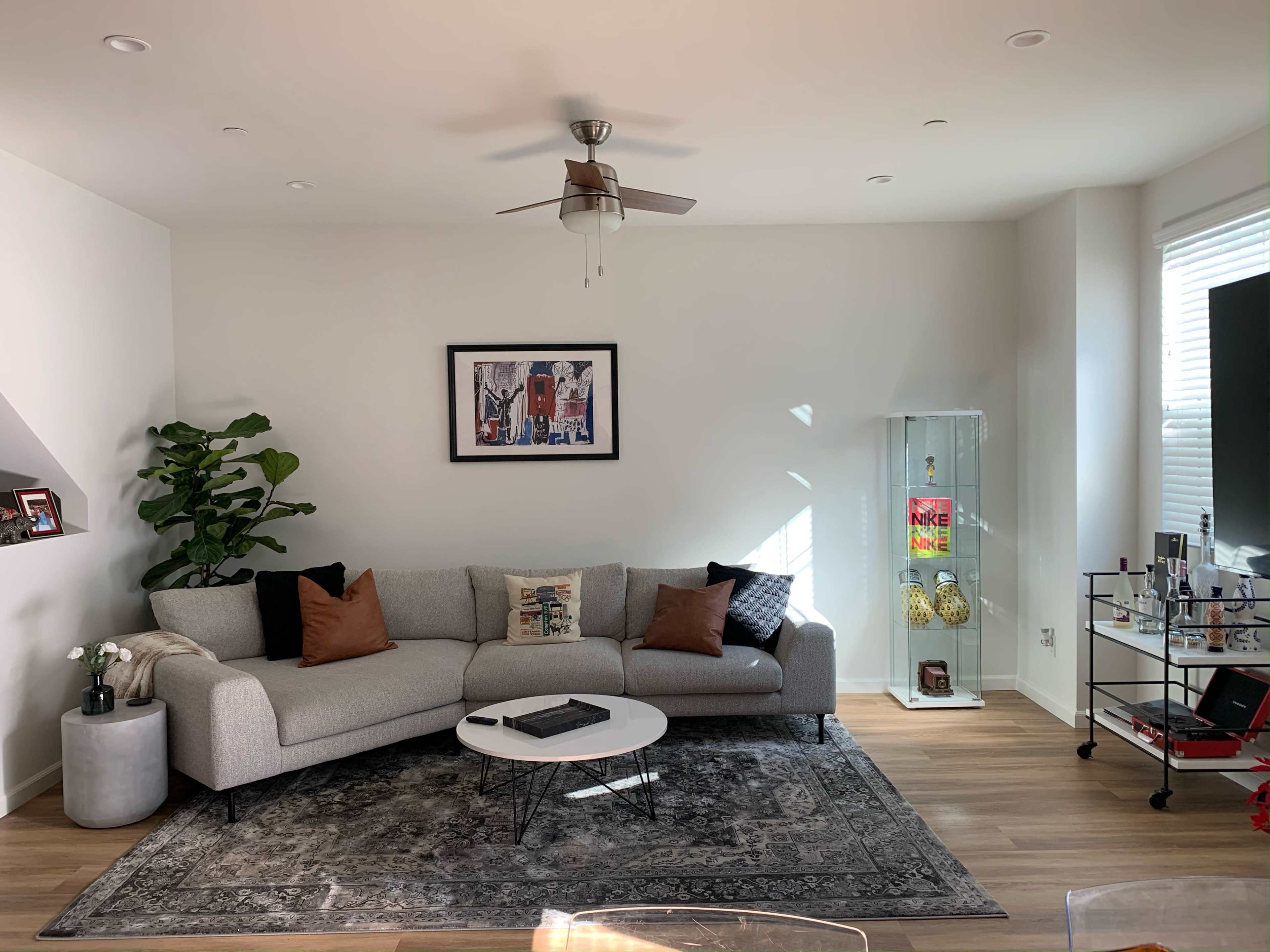The image depicts a modern living room featuring a gray sofa with decorative pillows, a round coffee table, a plant by the wall, a glass display cabinet, and a television on a wheeled stand.