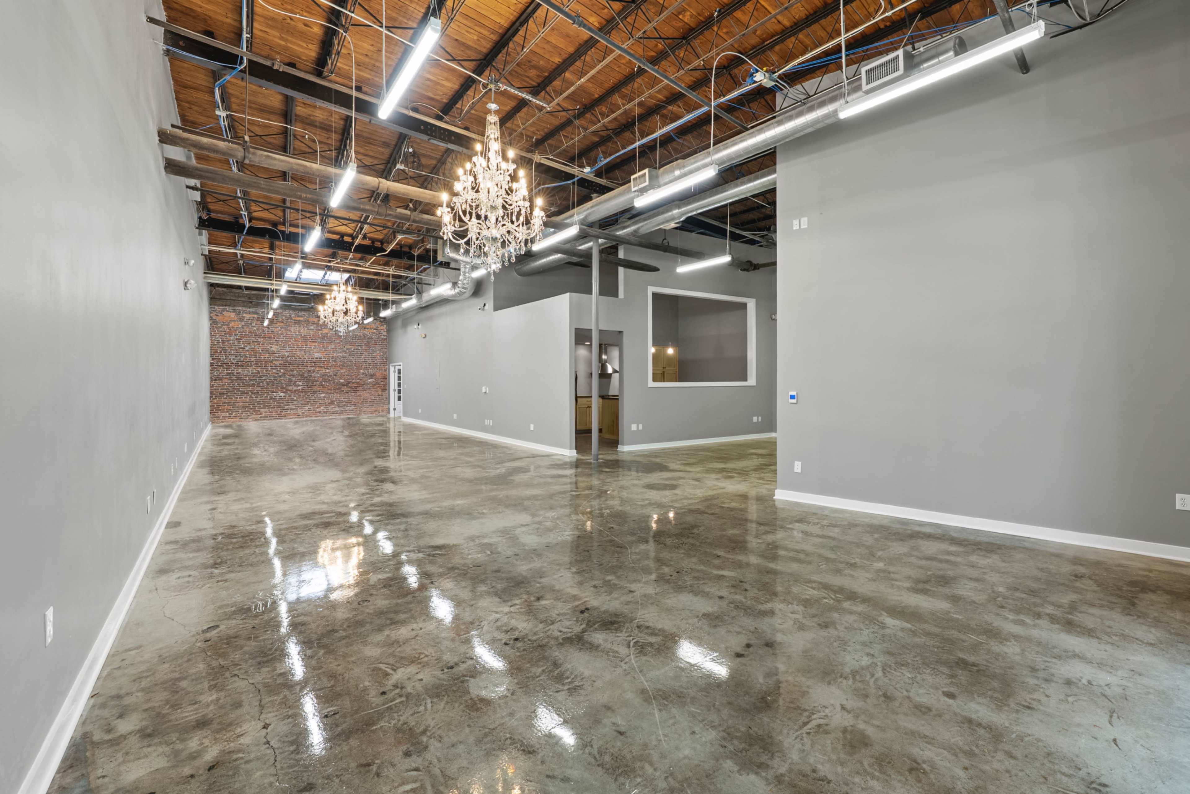 The image shows a spacious interior with polished concrete floors, exposed wooden beams, and two chandeliers hanging from the ceiling, alongside a partially visible brick wall.