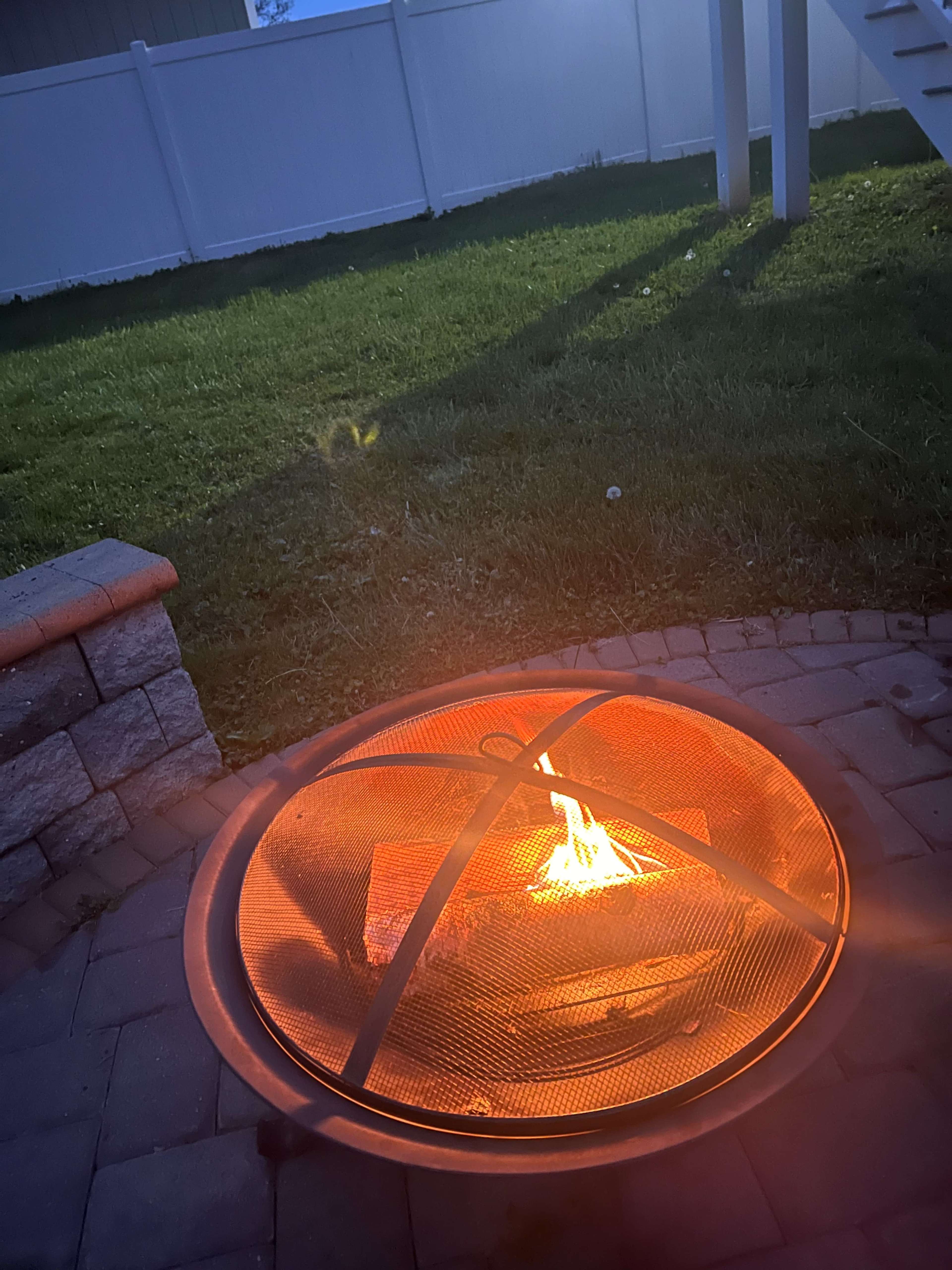 A fire burns in a circular fire pit, surrounded by stone pavers, in a grassy backyard with a white fence in the background.