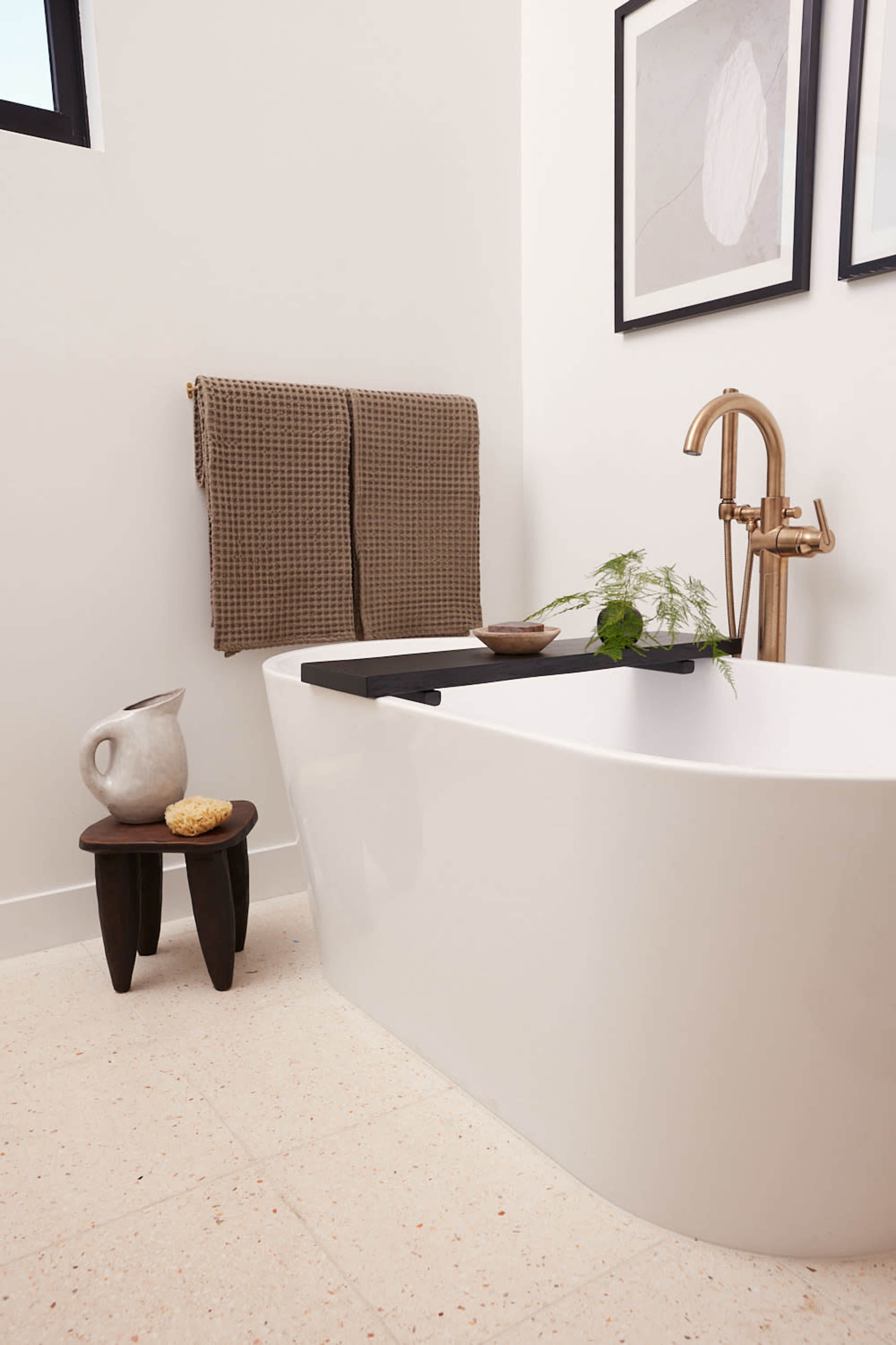 The image shows a modern bathroom featuring a freestanding white bathtub with a black accent tray, a small wooden stool, a decorative pitcher, and framed artwork on the wall.