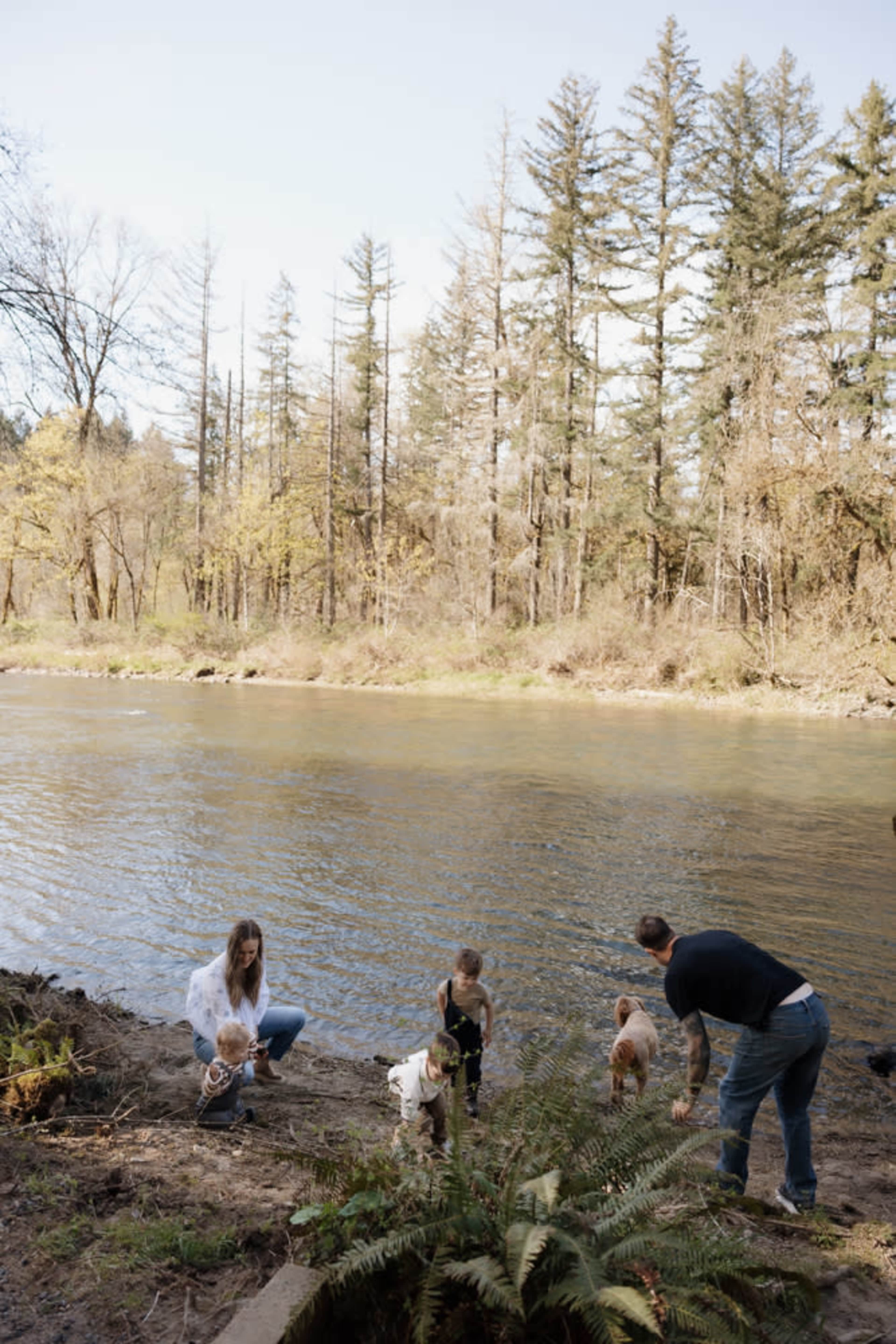A family with two children and two dogs is exploring the bank of a river surrounded by trees.