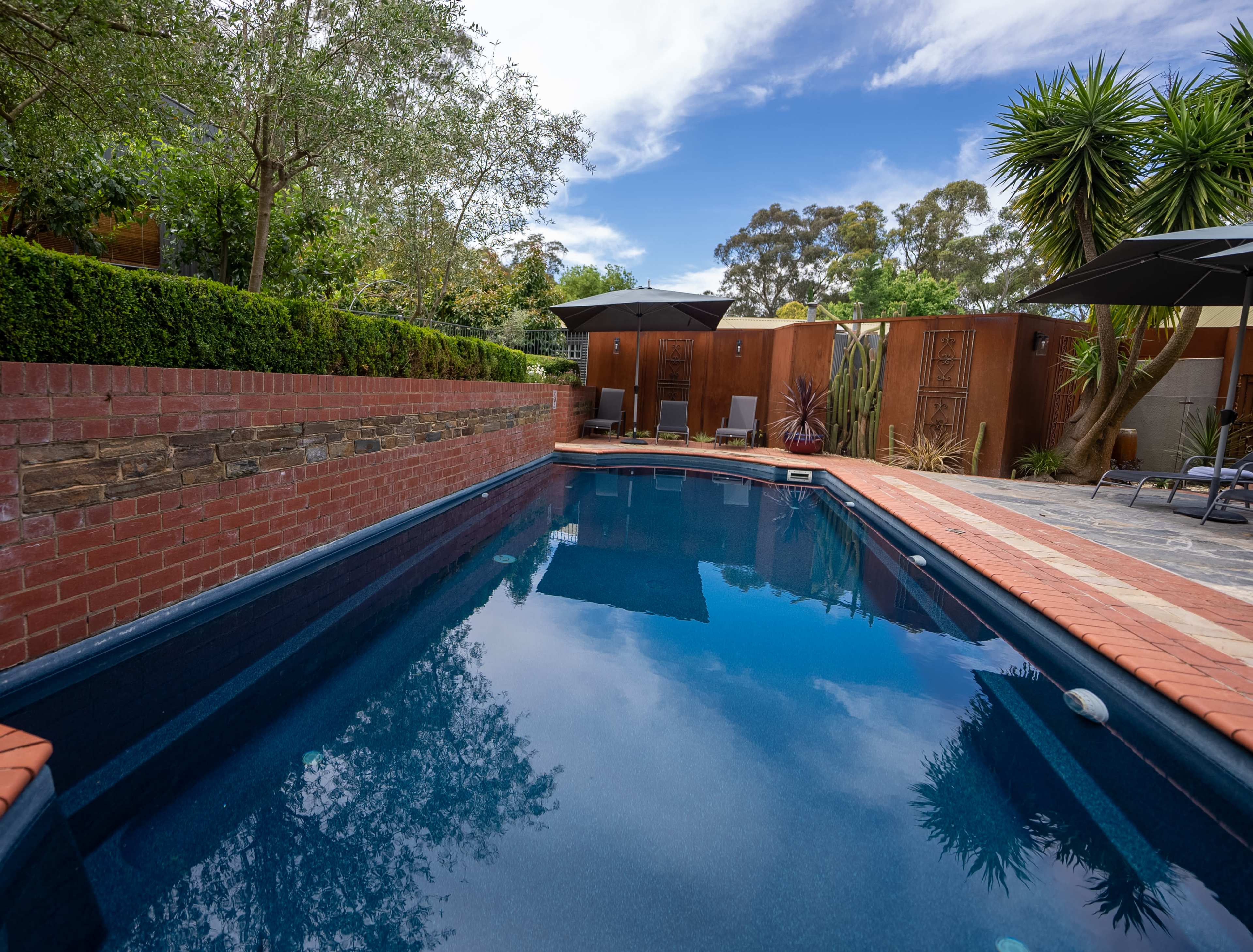 The image shows a rectangular swimming pool surrounded by stone and brick walls, with lounge chairs and umbrellas set up nearby under a clear blue sky.