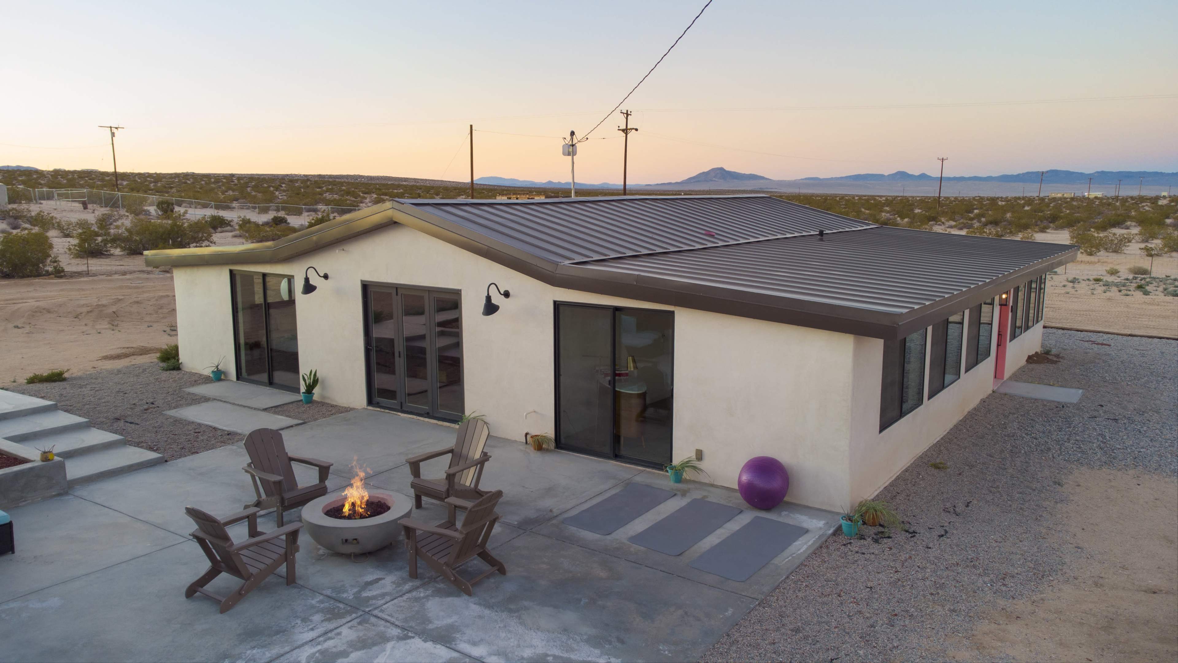 A modern house with a flat roof and large glass windows sits among desert landscaping, featuring an outdoor seating area with a fire pit.