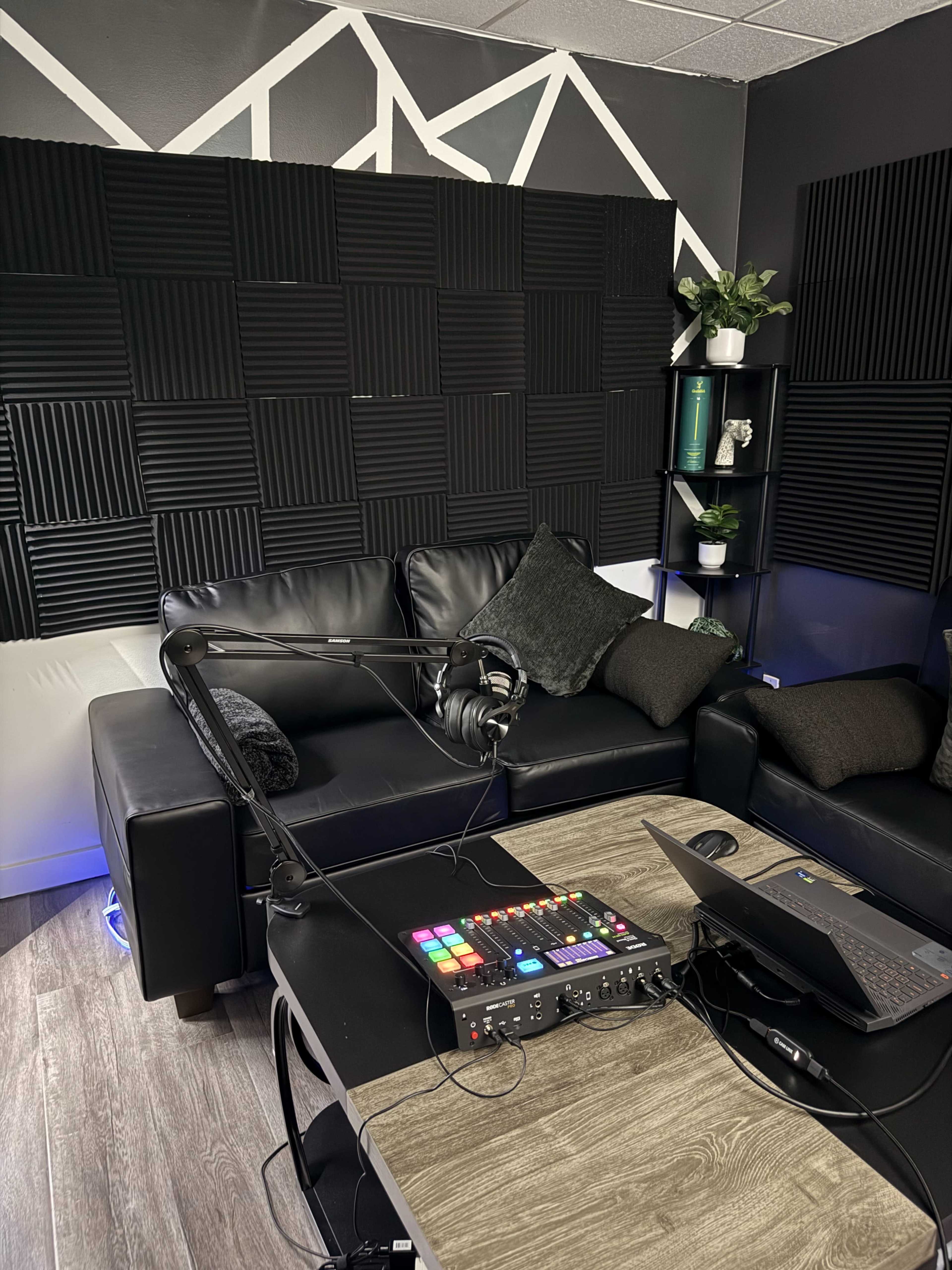The image shows a recording studio setup featuring a black leather couch, a desktop with a mixing console, a laptop, and soundproof foam panels on the walls.