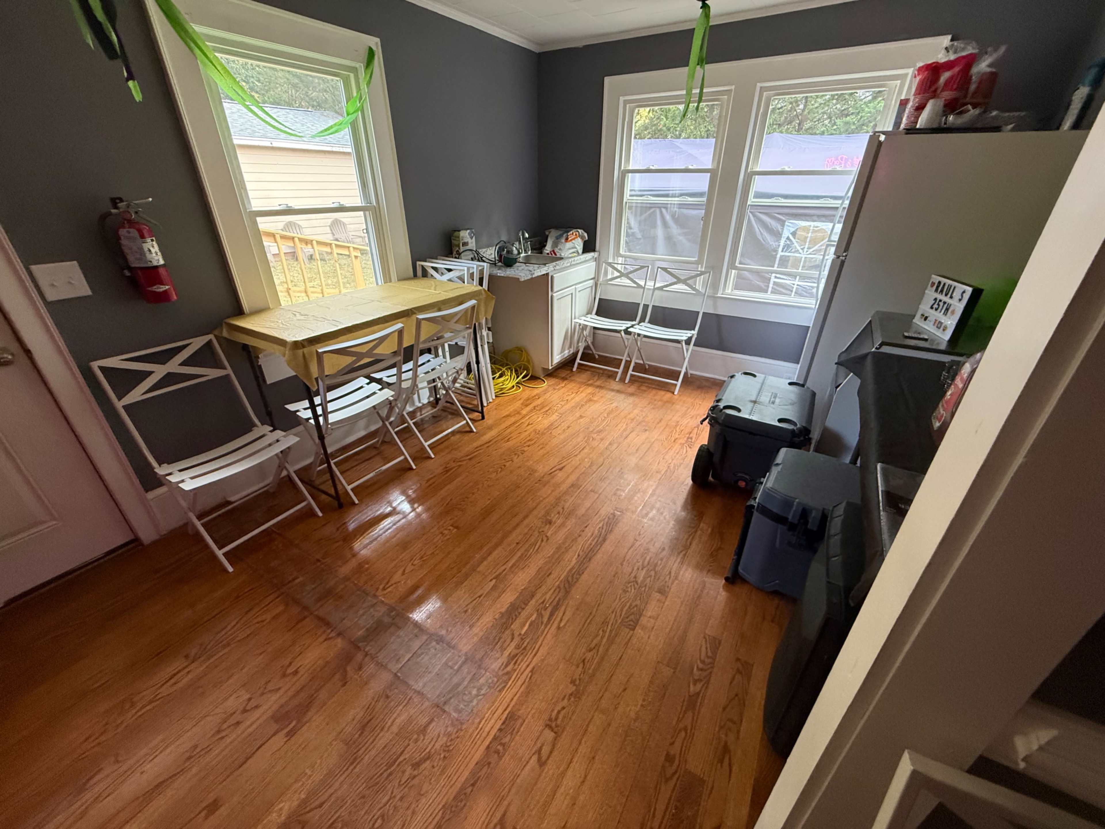 The image shows a room with a wooden floor, a small table with chairs, a refrigerator, and a window with light fabric curtains.