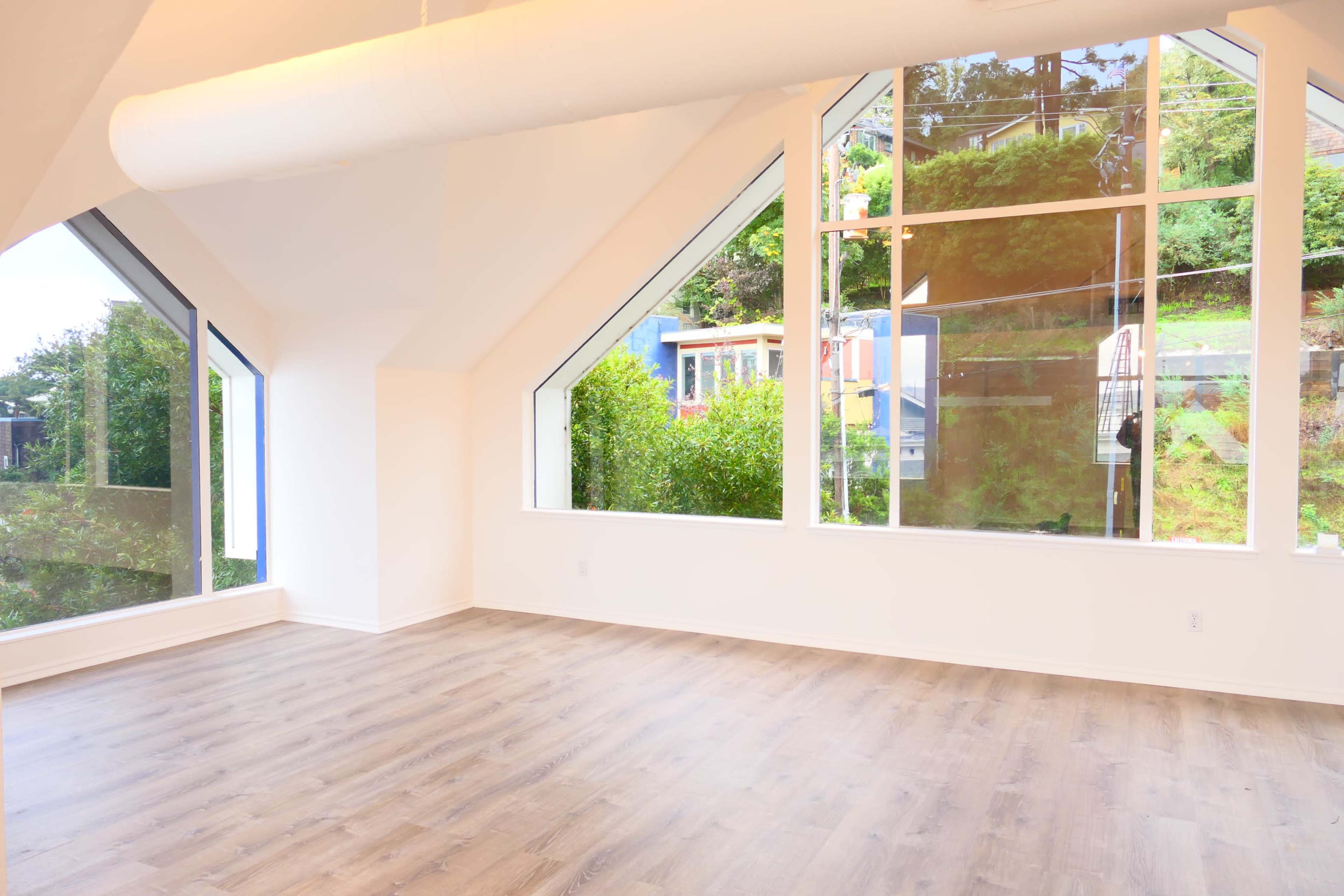 A spacious room with large triangular windows overlooking greenery, and a light-colored wood floor.