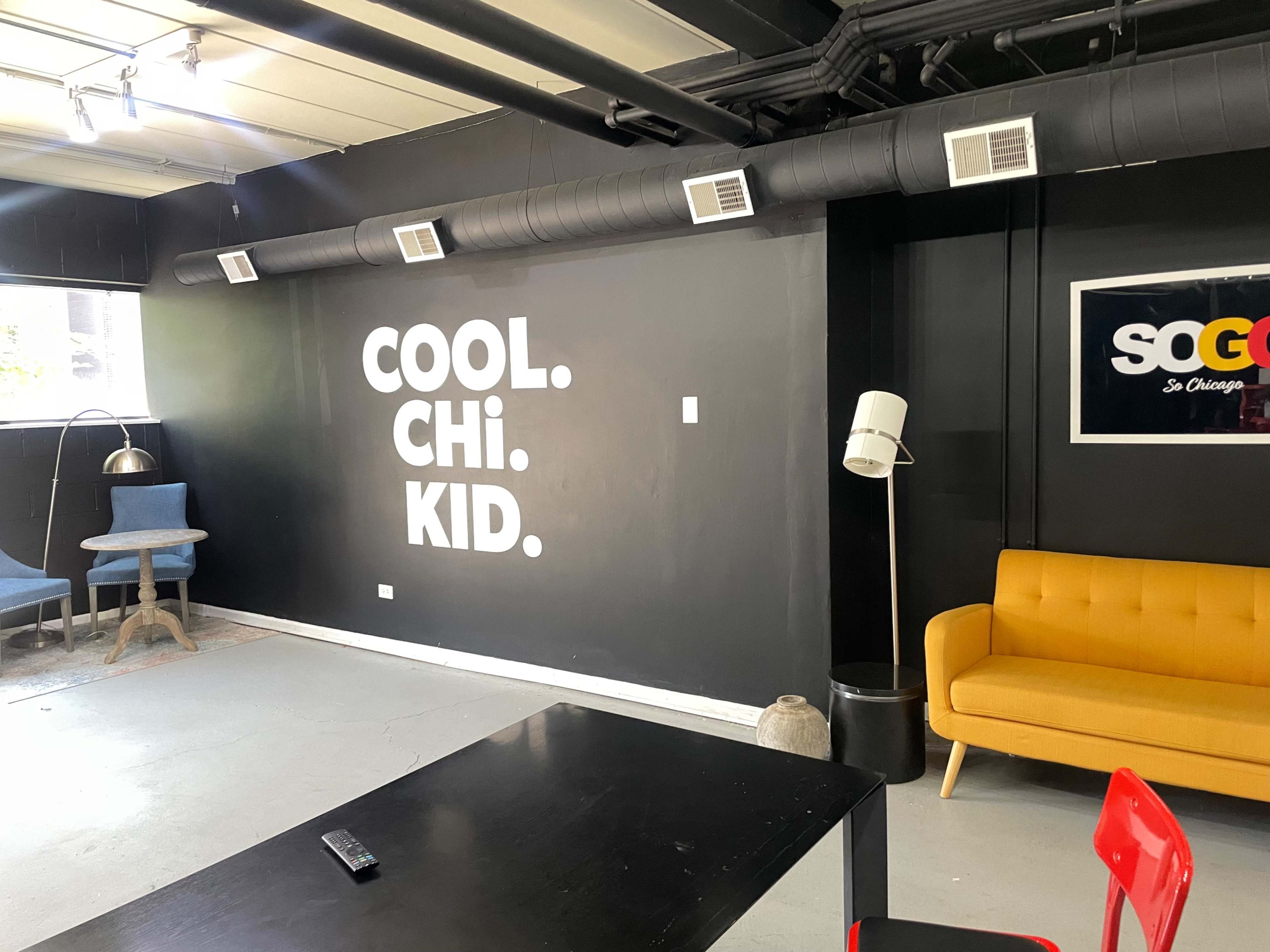 A modern interior with a black wall featuring the words "COOL. CHI. KID." in large white letters, a yellow couch, a blue chair, and a table.