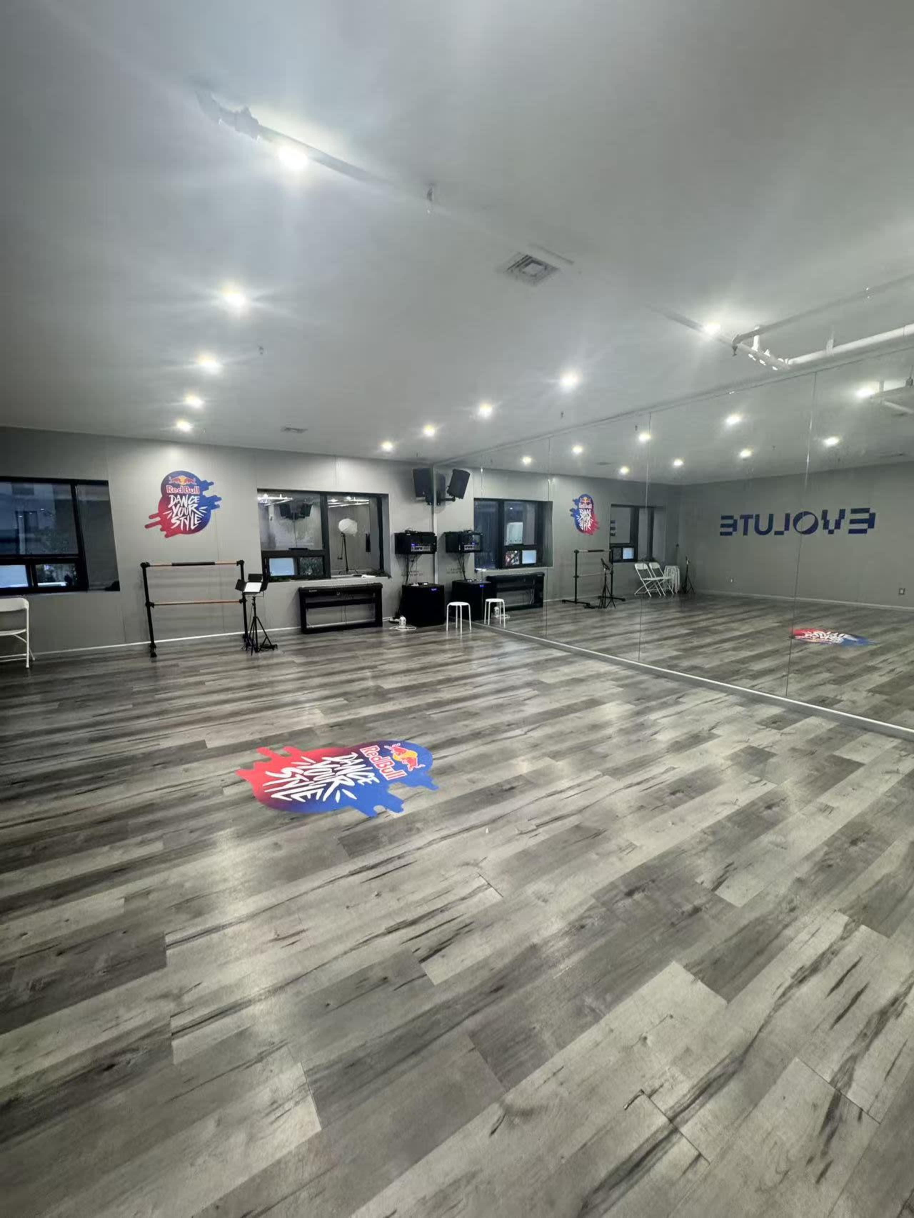 The image shows a spacious dance studio with wooden flooring, mirrors on one wall, and a logo on the floor.