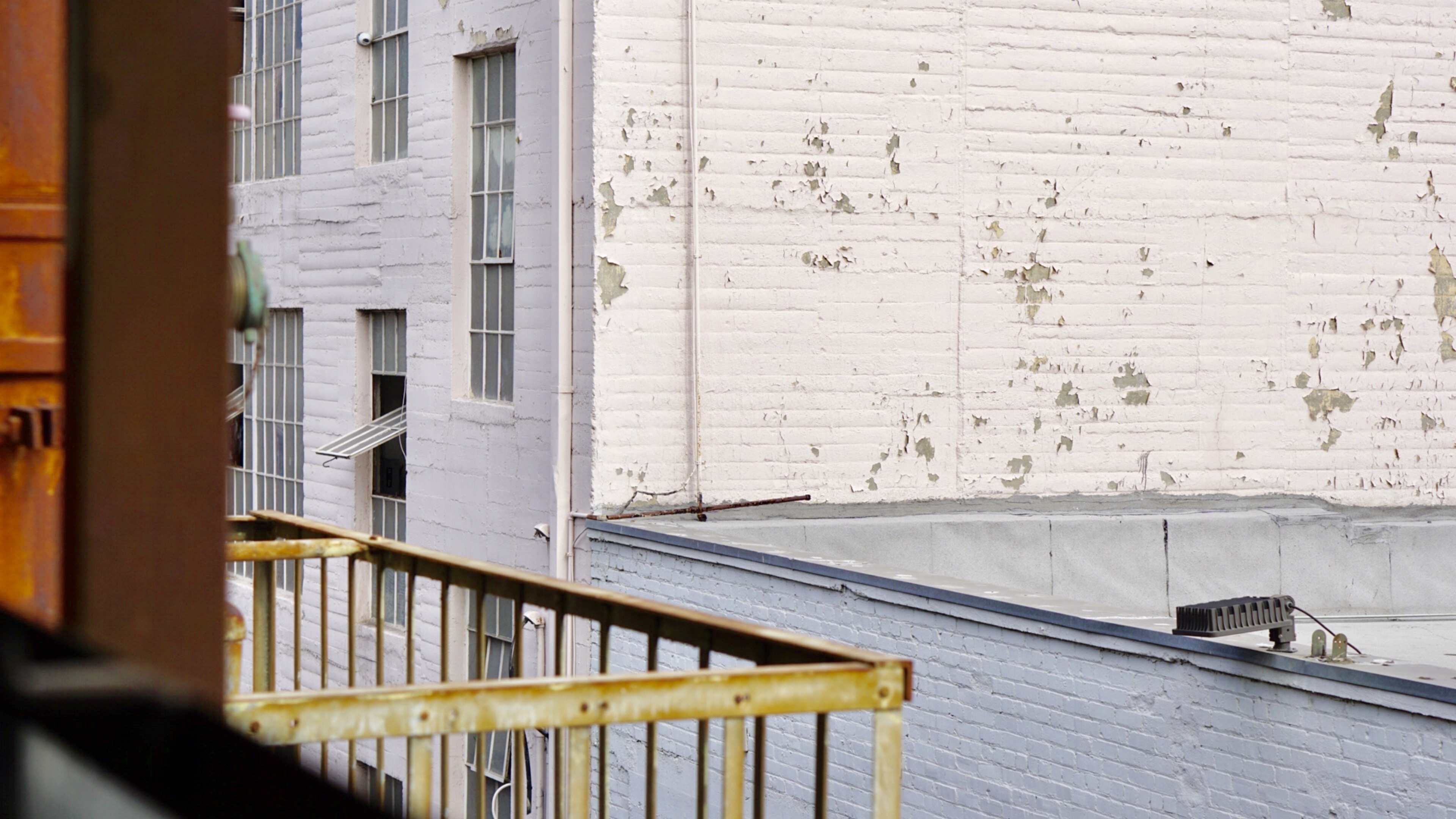The image shows a view of a weathered brick wall with visible paint peeling, alongside a metal balcony railing.