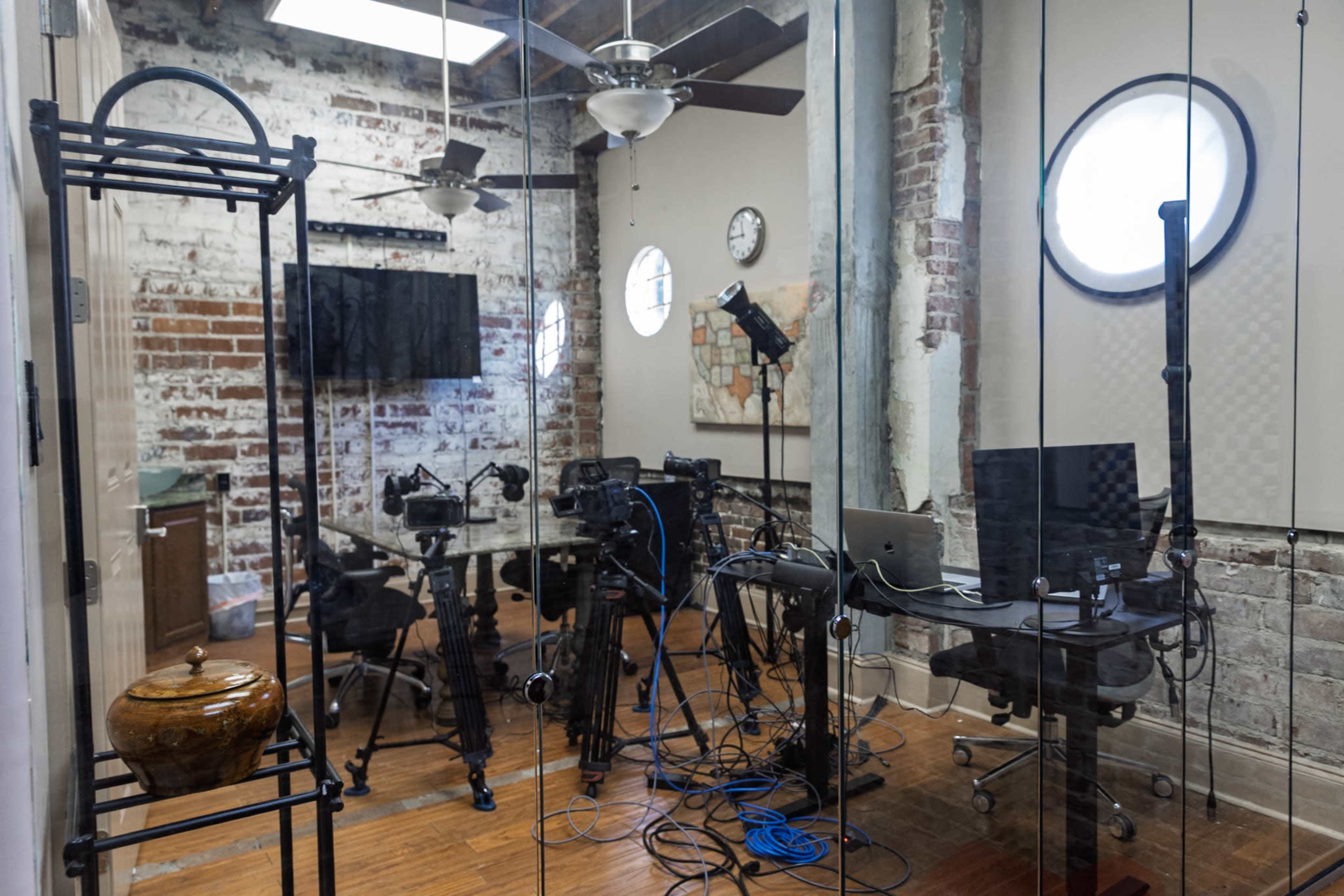 The image shows a modern conference room with exposed brick walls, a large glass table surrounded by chairs, and various equipment set up for a meeting or video recording.