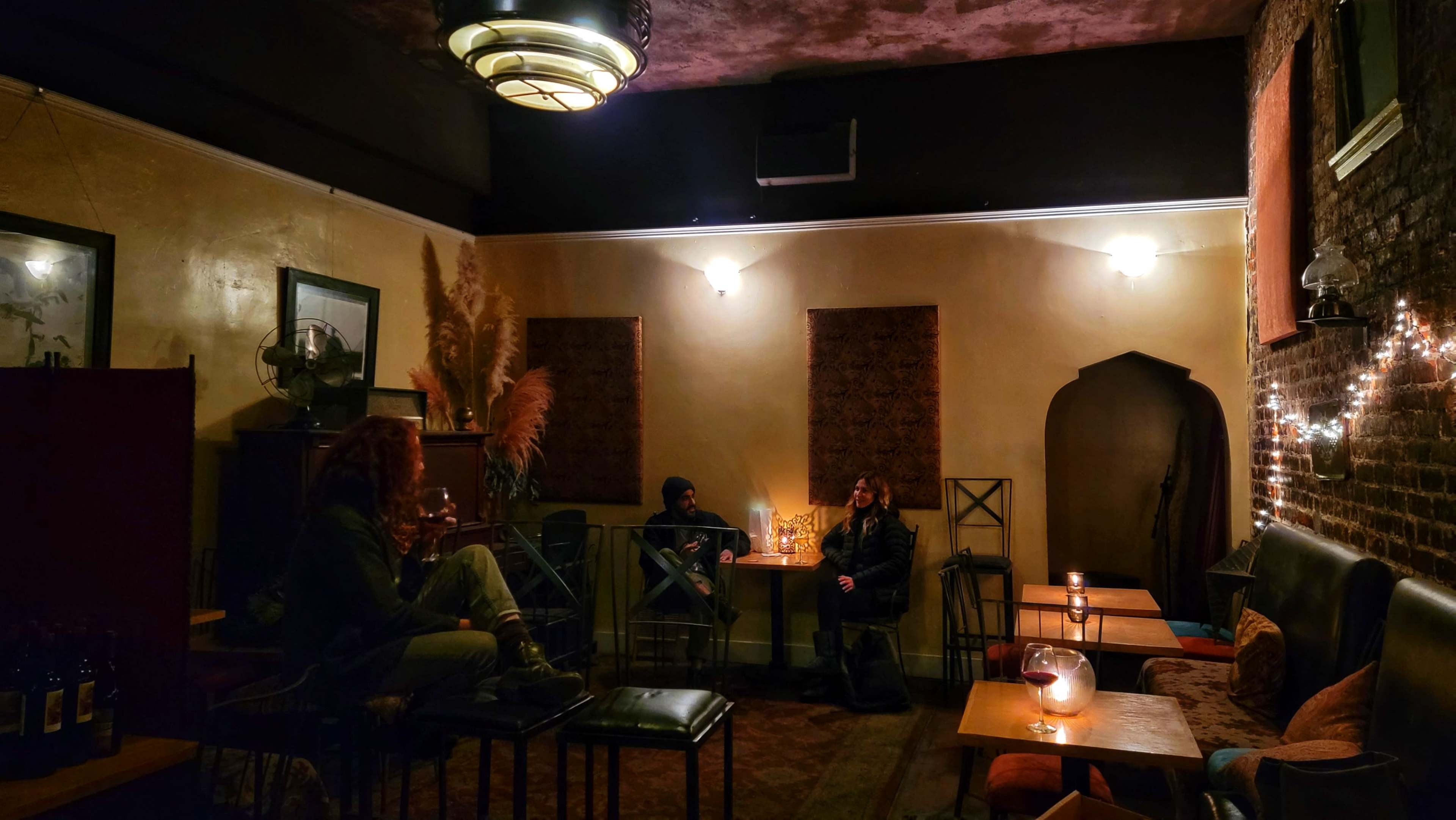 A cozy interior of a dimly lit lounge features two seated patrons among tables with candles and decorative elements.