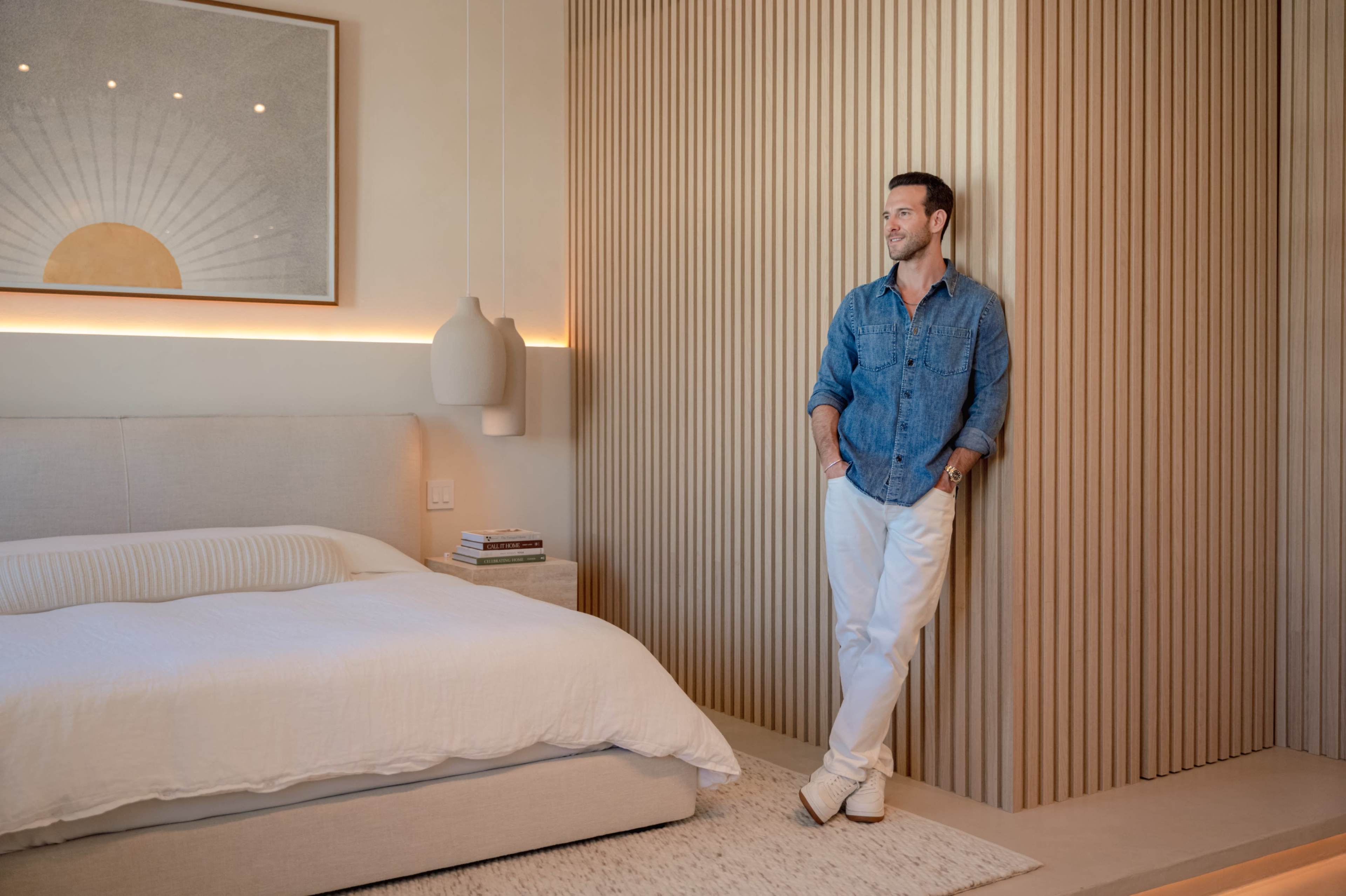 A man stands against a wooden accent wall in a modern bedroom, wearing a denim shirt and white trousers, with a bed and bedside table in the background.