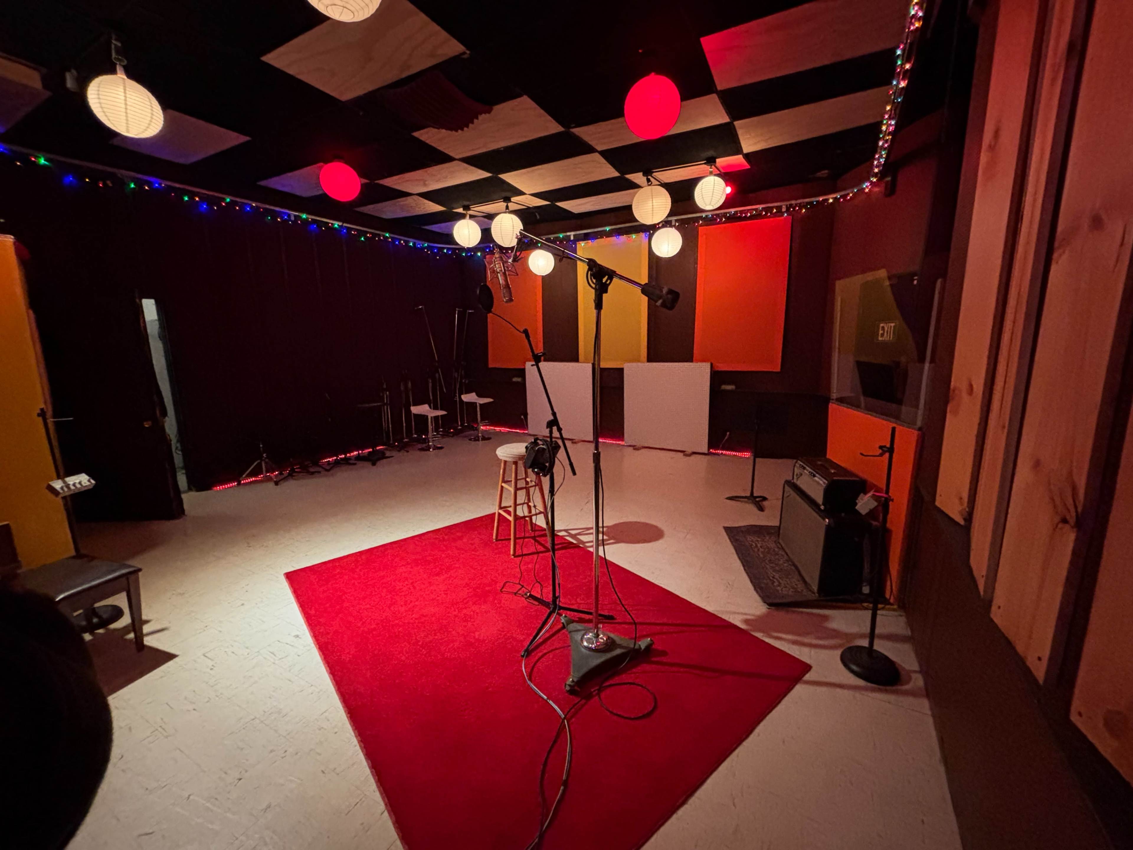 The image shows a recording studio with various microphones set up on a red carpet, surrounded by colorful lighting and soundproofing panels.