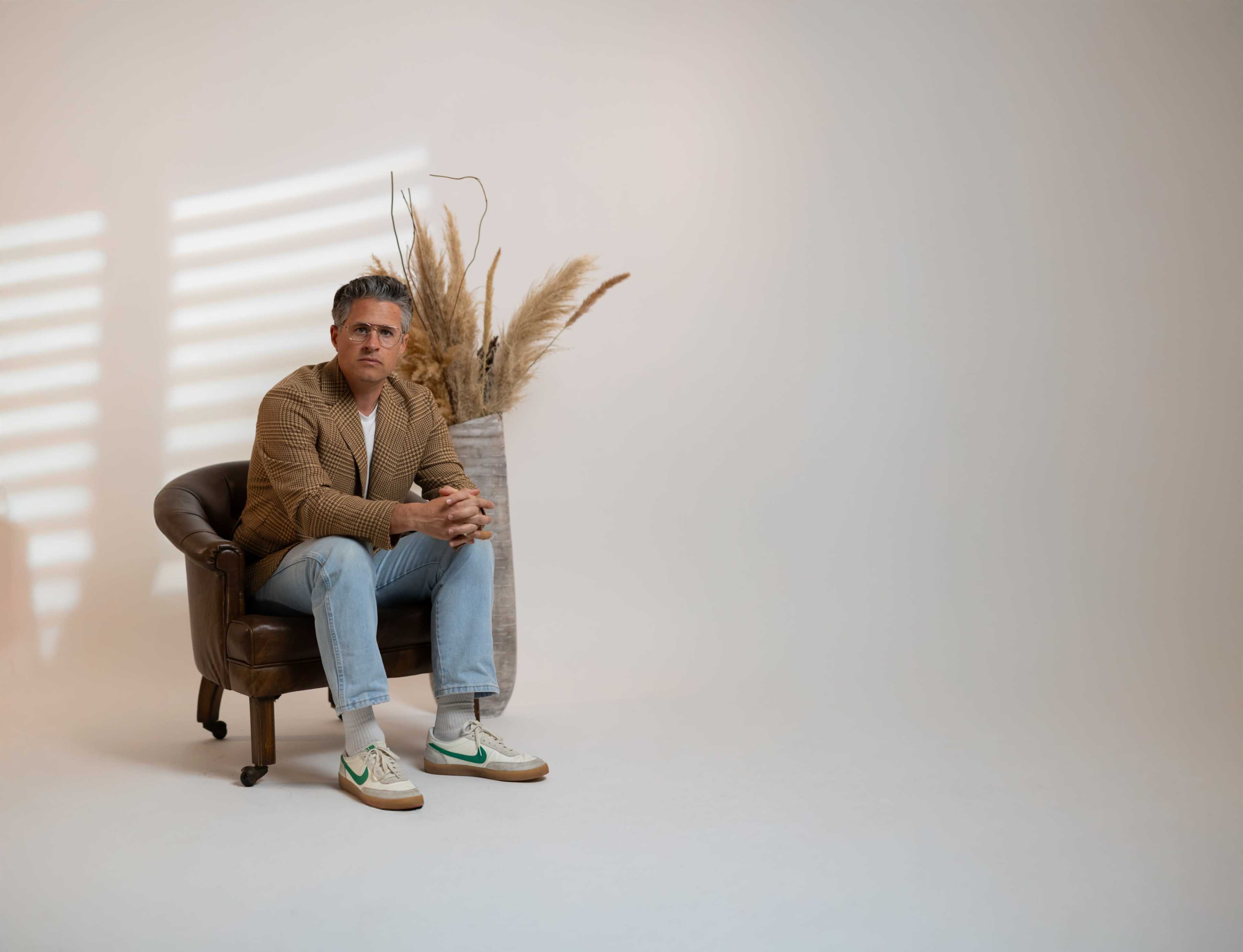 A man sits in a brown leather armchair, wearing a beige cardigan and light blue jeans, with decorative pampas grass in a vase beside him.