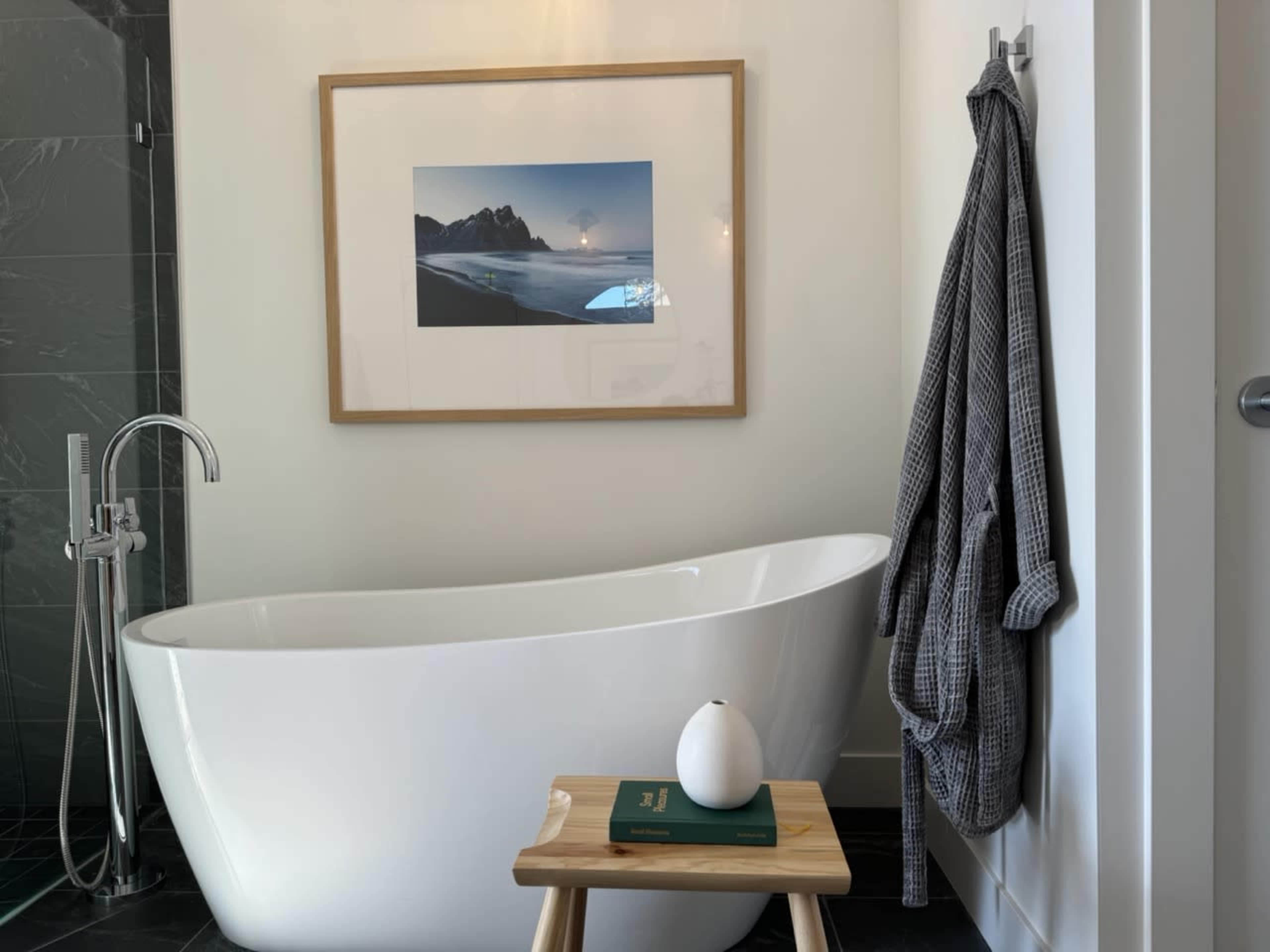 A freestanding white bathtub is positioned next to a framed landscape photograph, accompanied by a wooden stool holding a decorative item and a book.