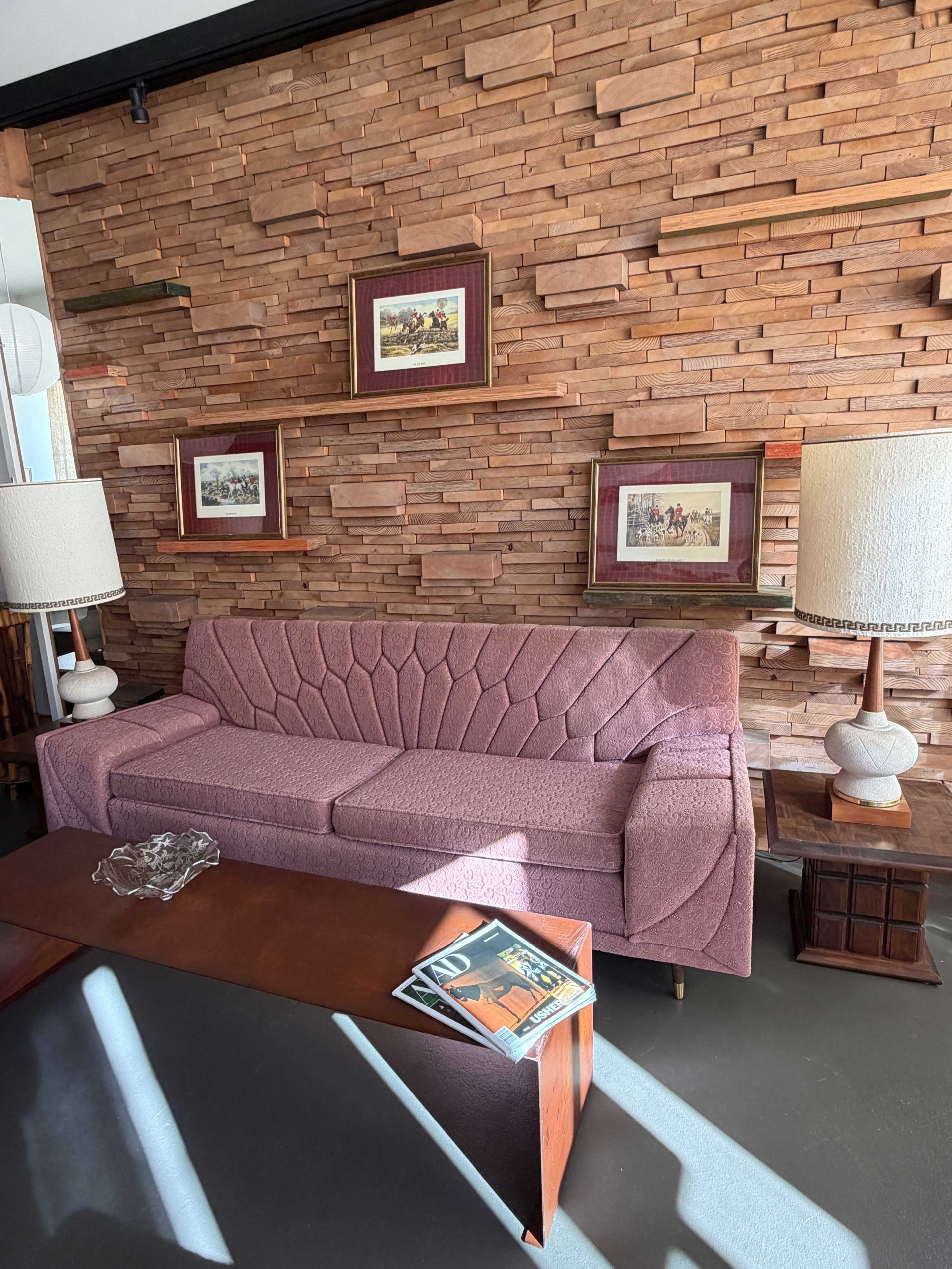 The image shows a mid-century modern living room with a pink sofa, wooden accent wall, two lamps, and a low coffee table displaying magazines and an ashtray.
