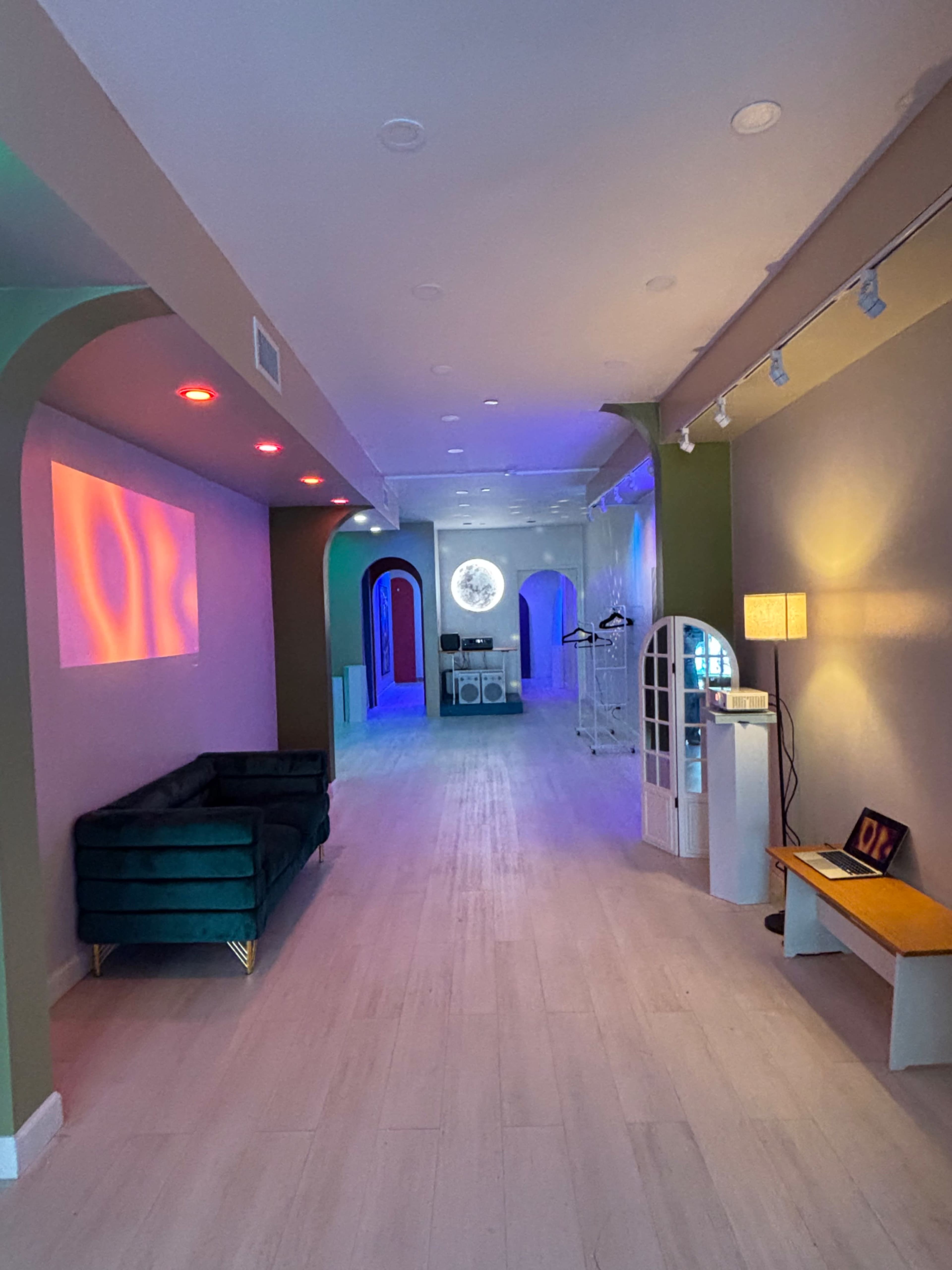 The image shows a modern interior space with arched doorways, colorful lighting, and a long hallway leading to a circular mirror at the end.