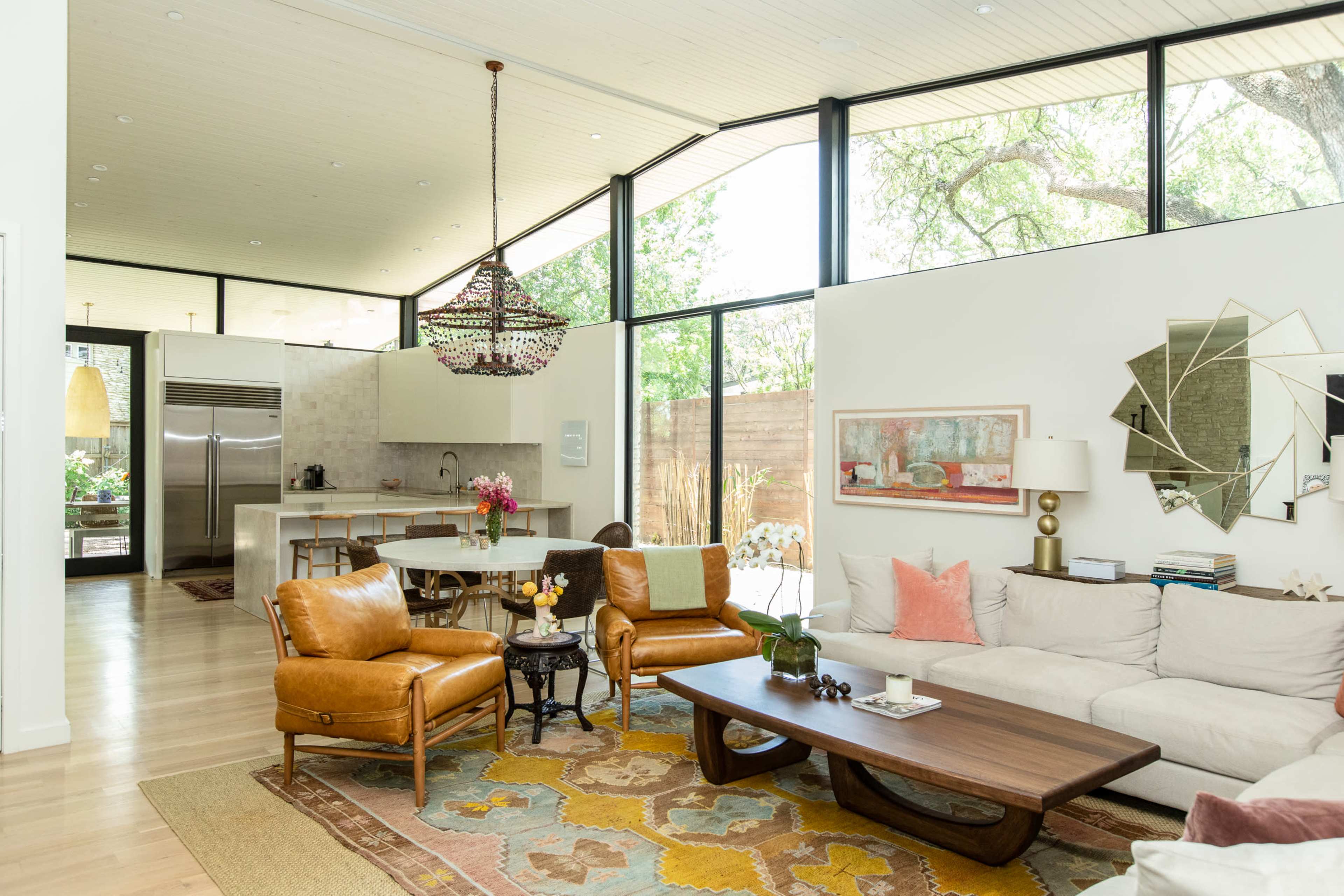 A modern living room with large windows allowing natural light, featuring brown leather chairs, a beige sofa, a wooden coffee table, and a decorative rug.