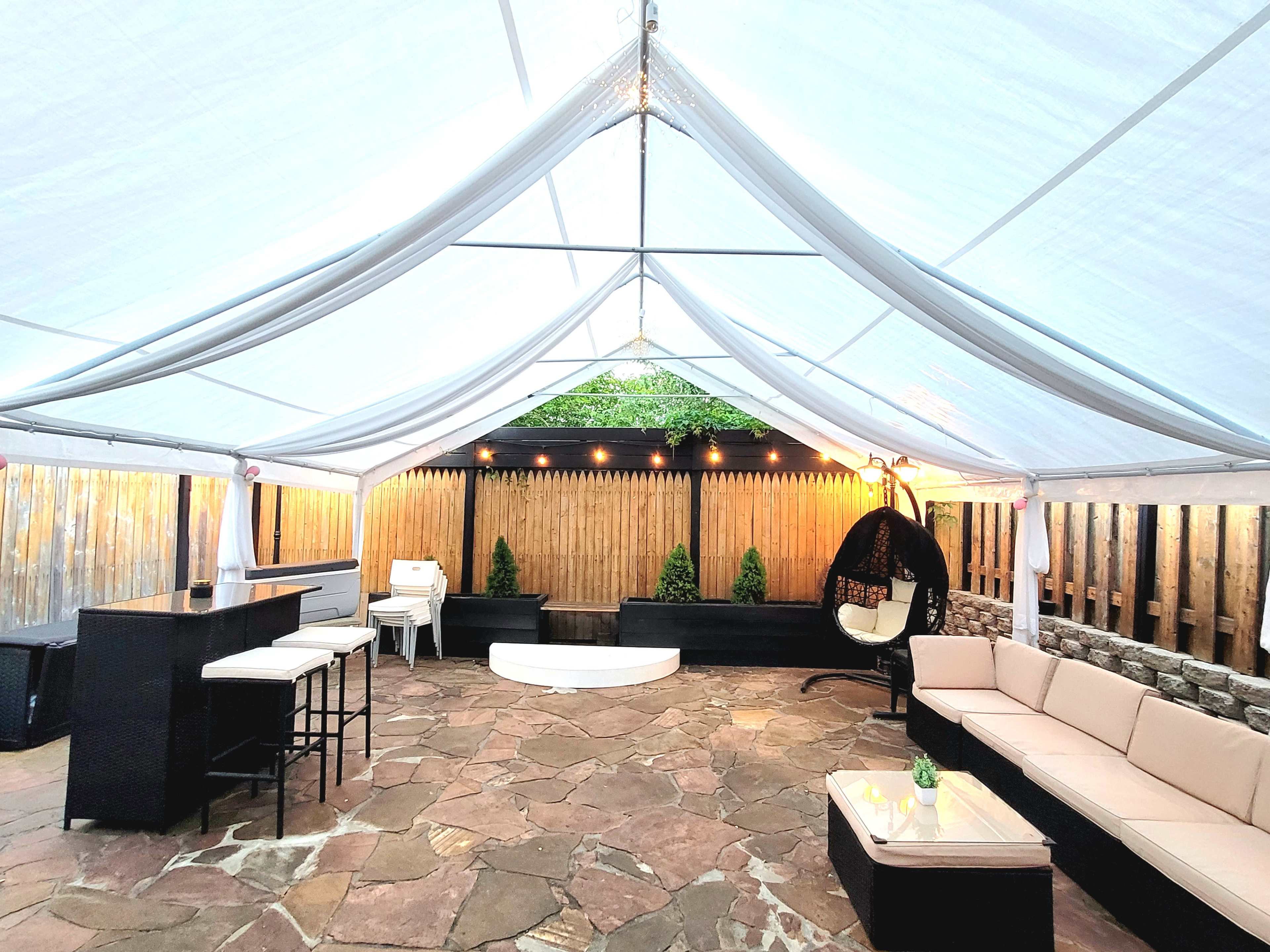 A tented outdoor patio features a stone floor, a sectional sofa, a hanging egg chair, a bar area, and potted plants against a wooden fence.