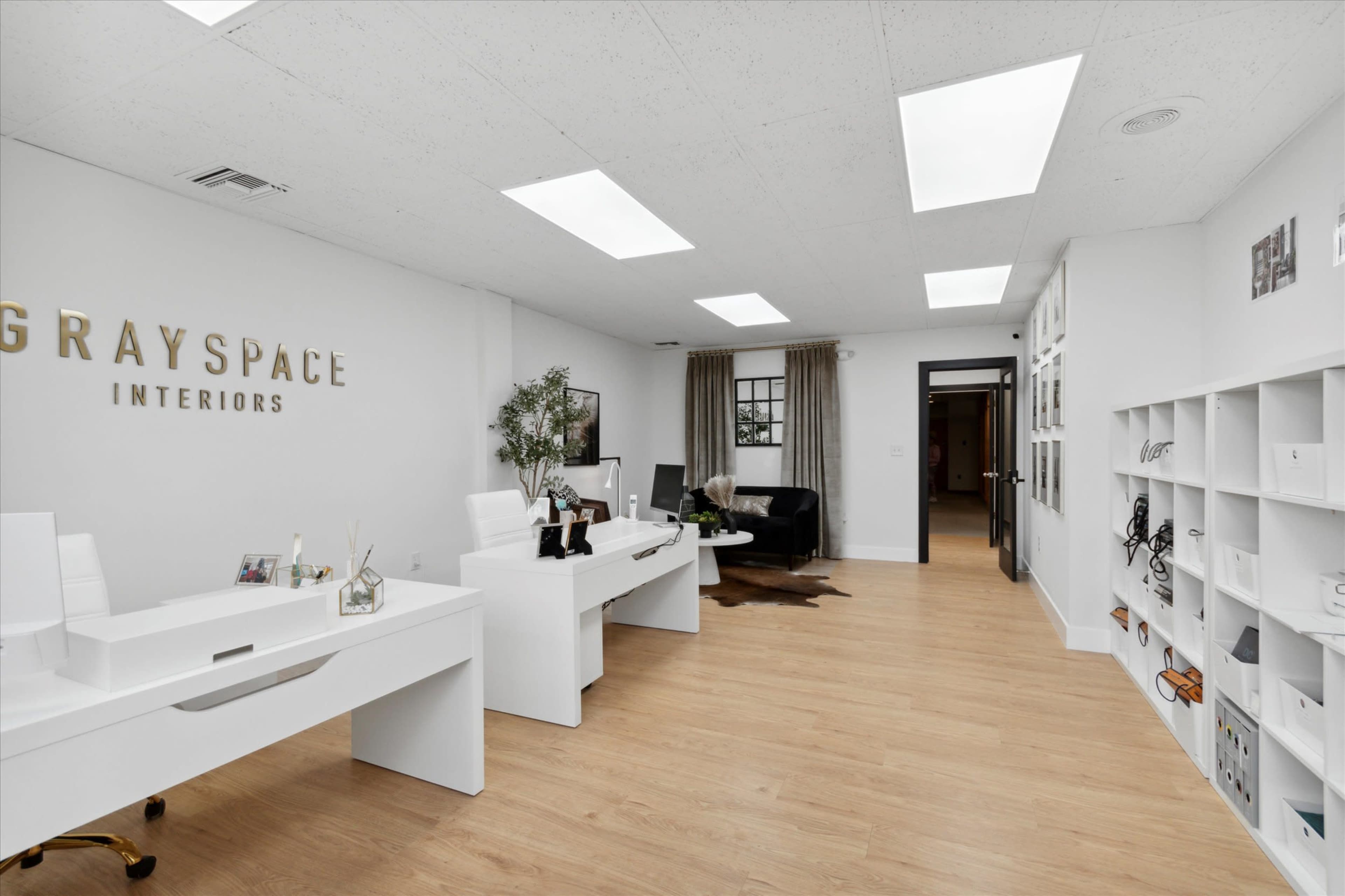 The image shows a modern office space featuring two white desks, a black seating area, and large skylights illuminating the room.