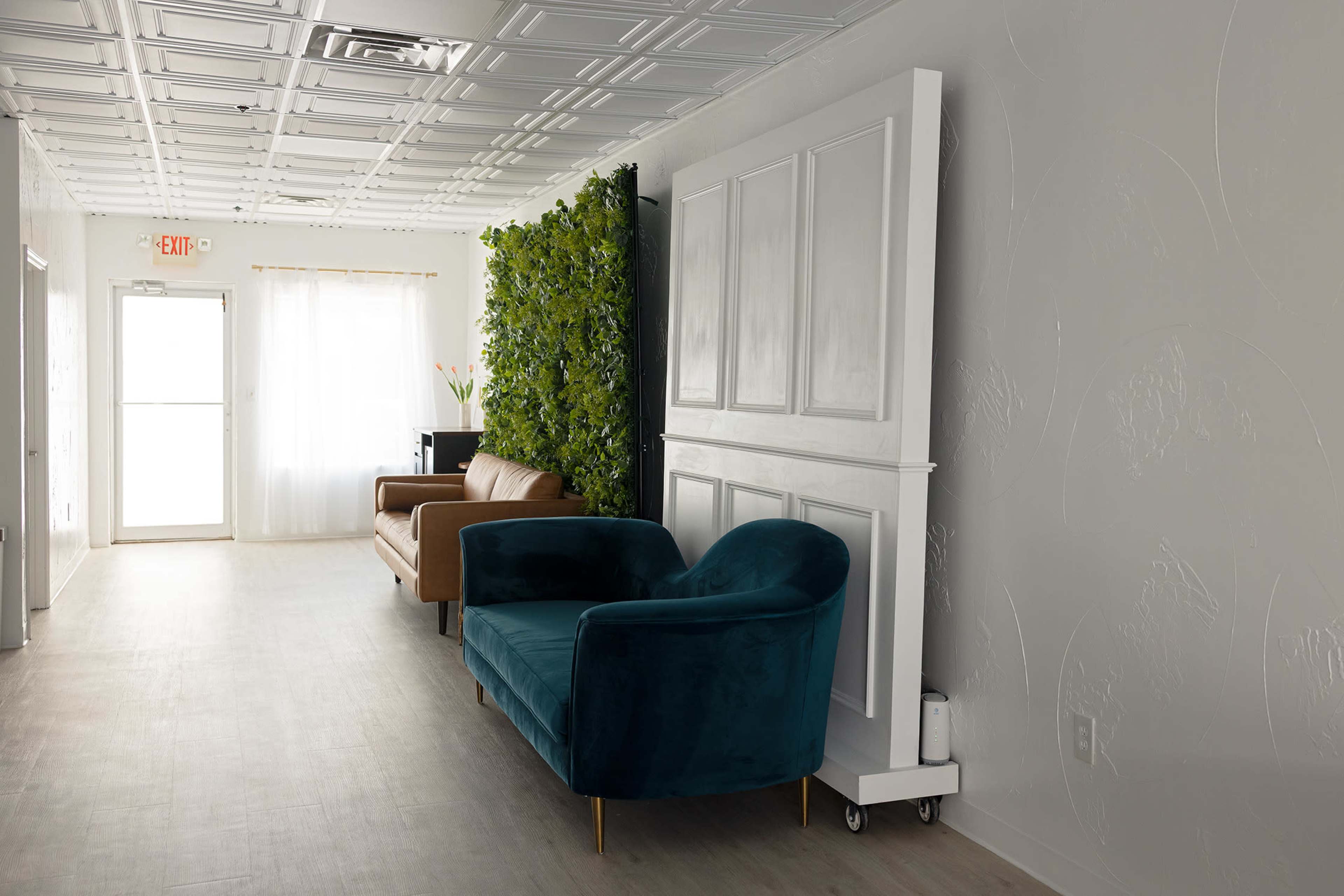 The image shows a hallway with two sofas, one teal and one brown, alongside a green vertical garden and a large white folding partition.