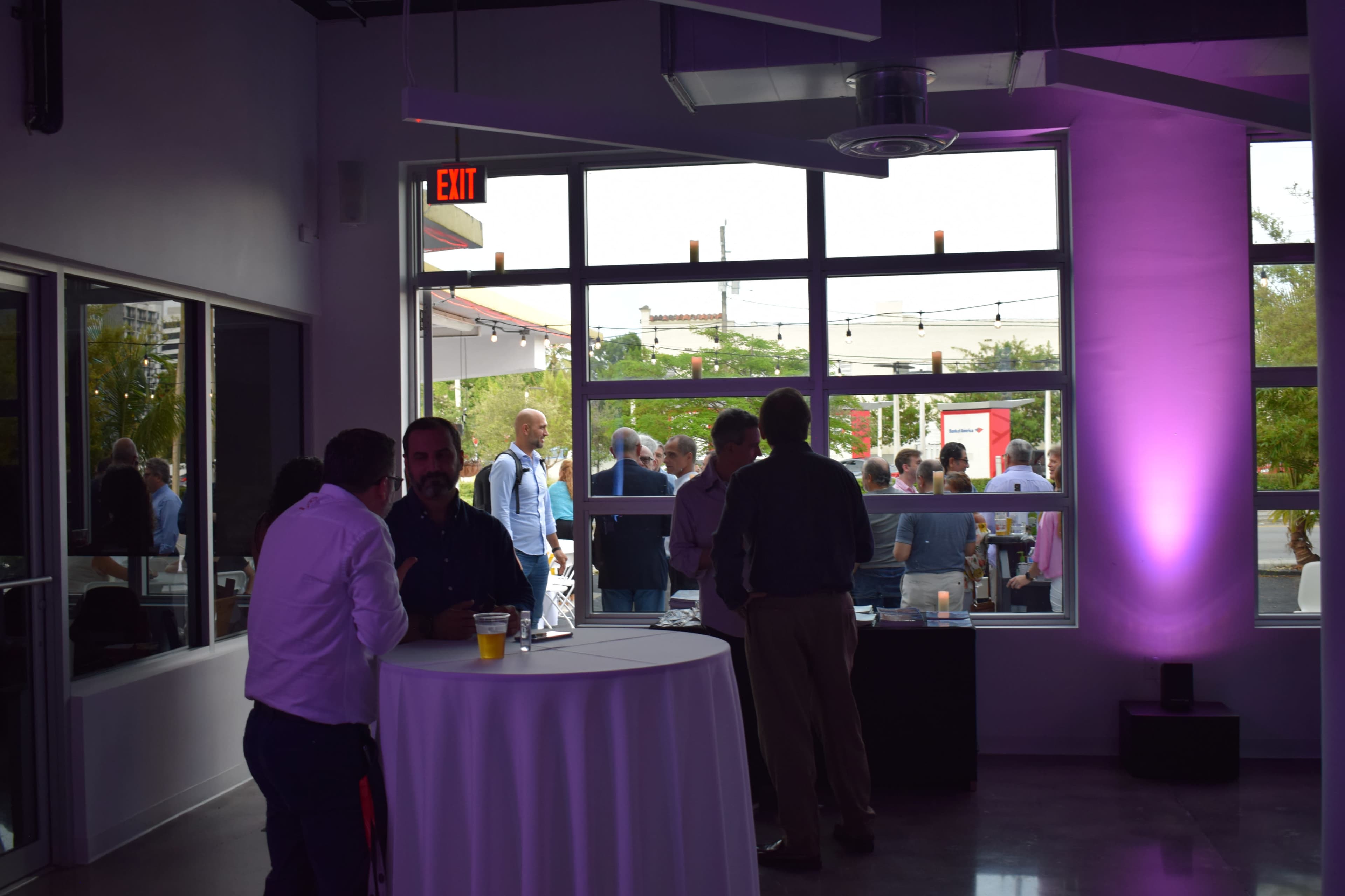 A group of people network in a brightly lit room with large windows overlooking an outdoor gathering.