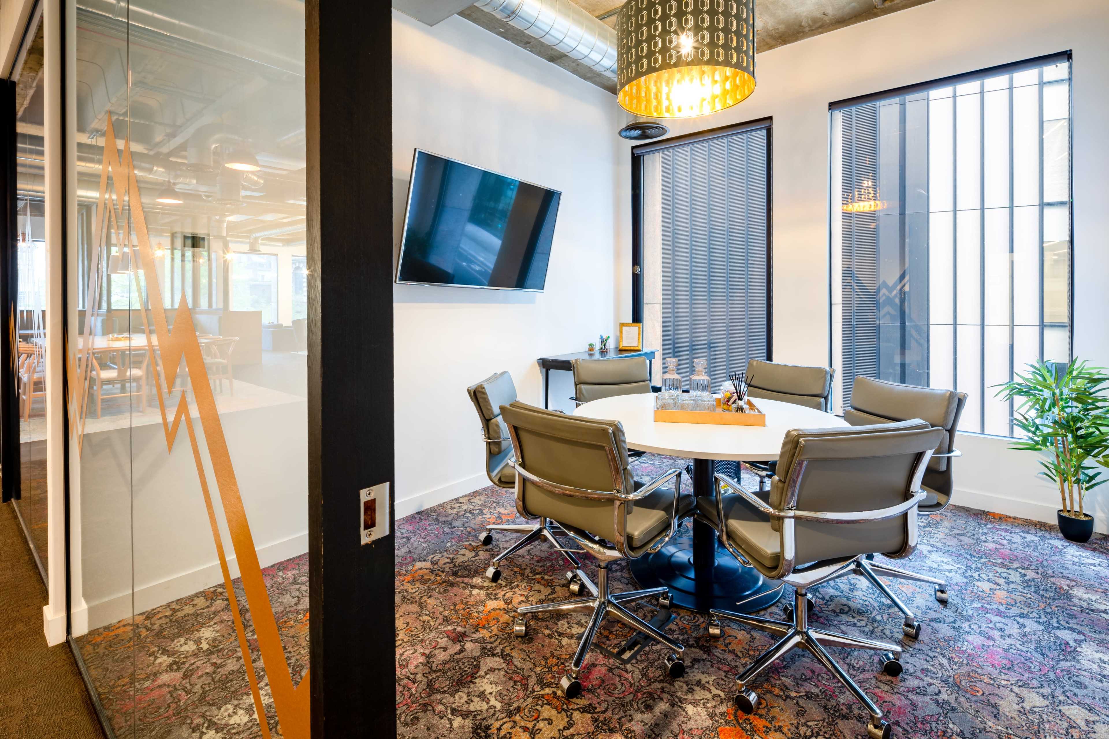 The image shows a modern conference room featuring a round table surrounded by ergonomic chairs, a wall-mounted television, and large windows with blinds.